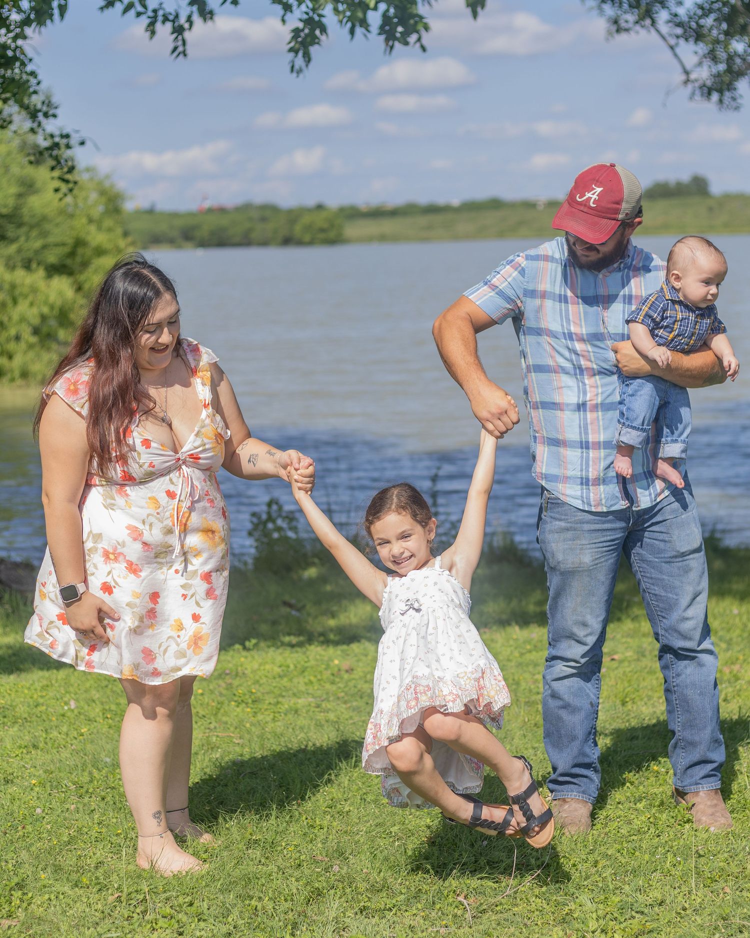 Playful family swings young girl by her hands near a scenic lakeside on a bright sunny day.