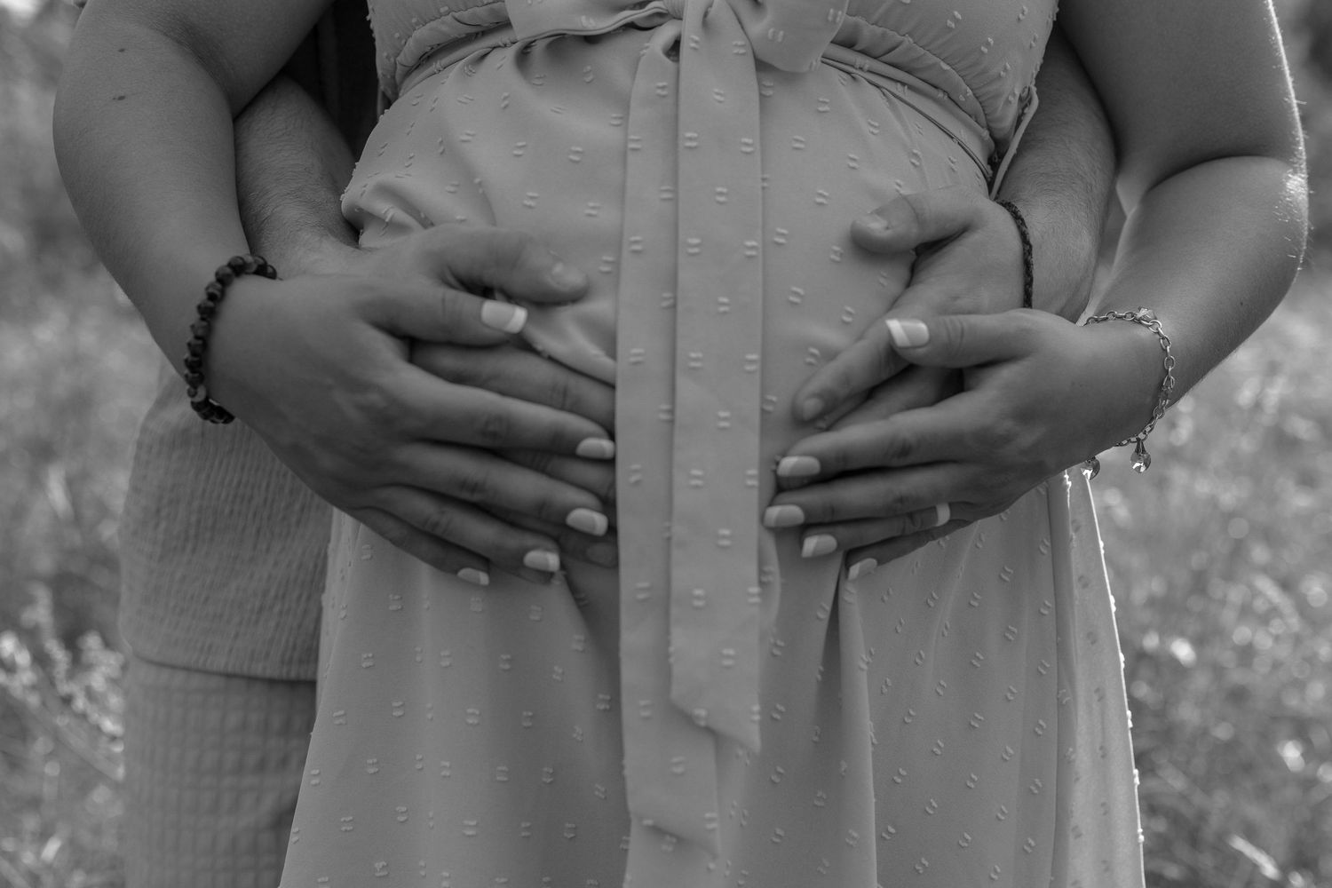 Black and white maternity photo of couple cradling pregnant belly with both sets of hands outdoors.
