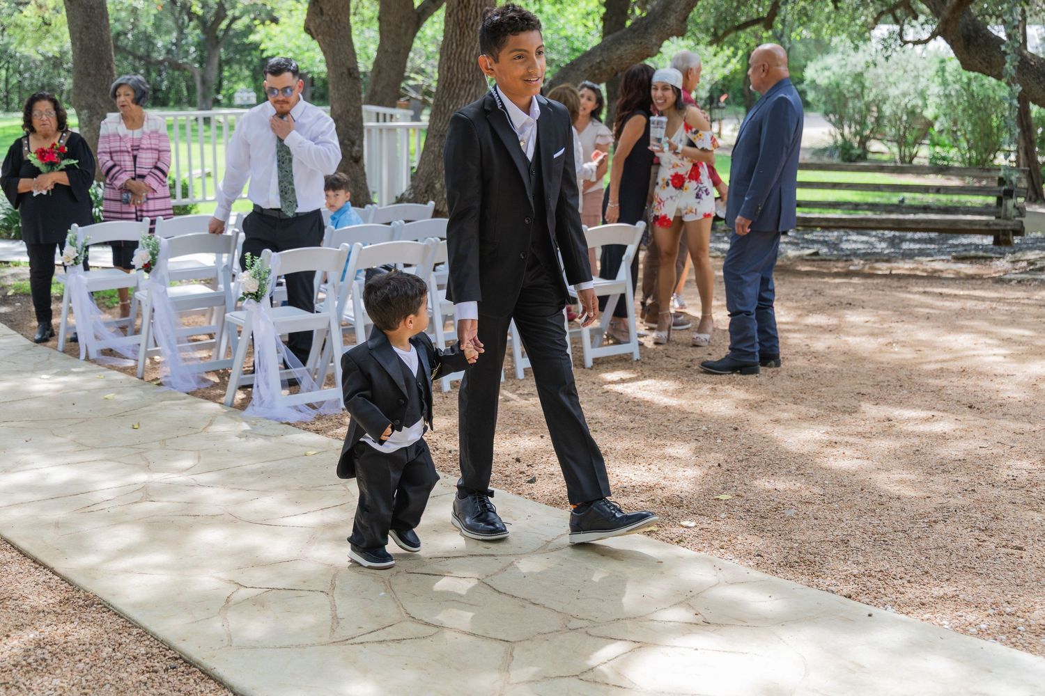 Young boy in tuxedo walks with older teen down wedding aisle at outdoor ceremony with seated guests.