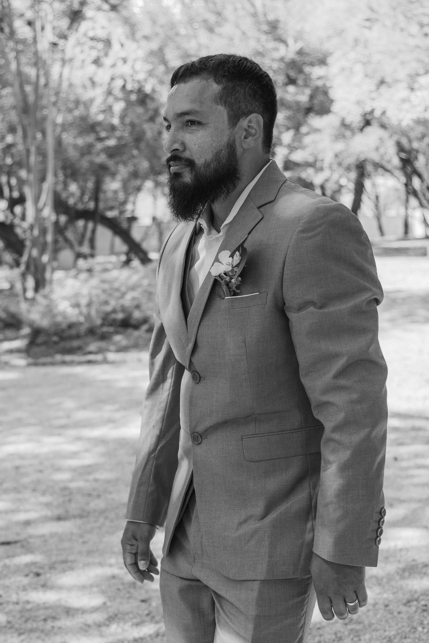 Groom in gray suit with boutonniere poses outdoors in black and white wedding portrait.