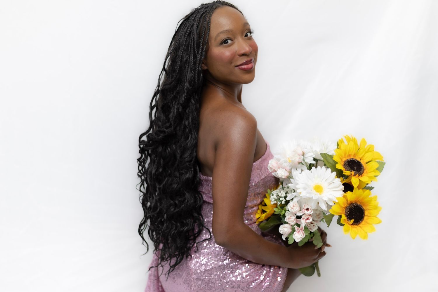 Smiling young Black woman in pink sequin dress holding sunflower and daisy bouquet against white background.