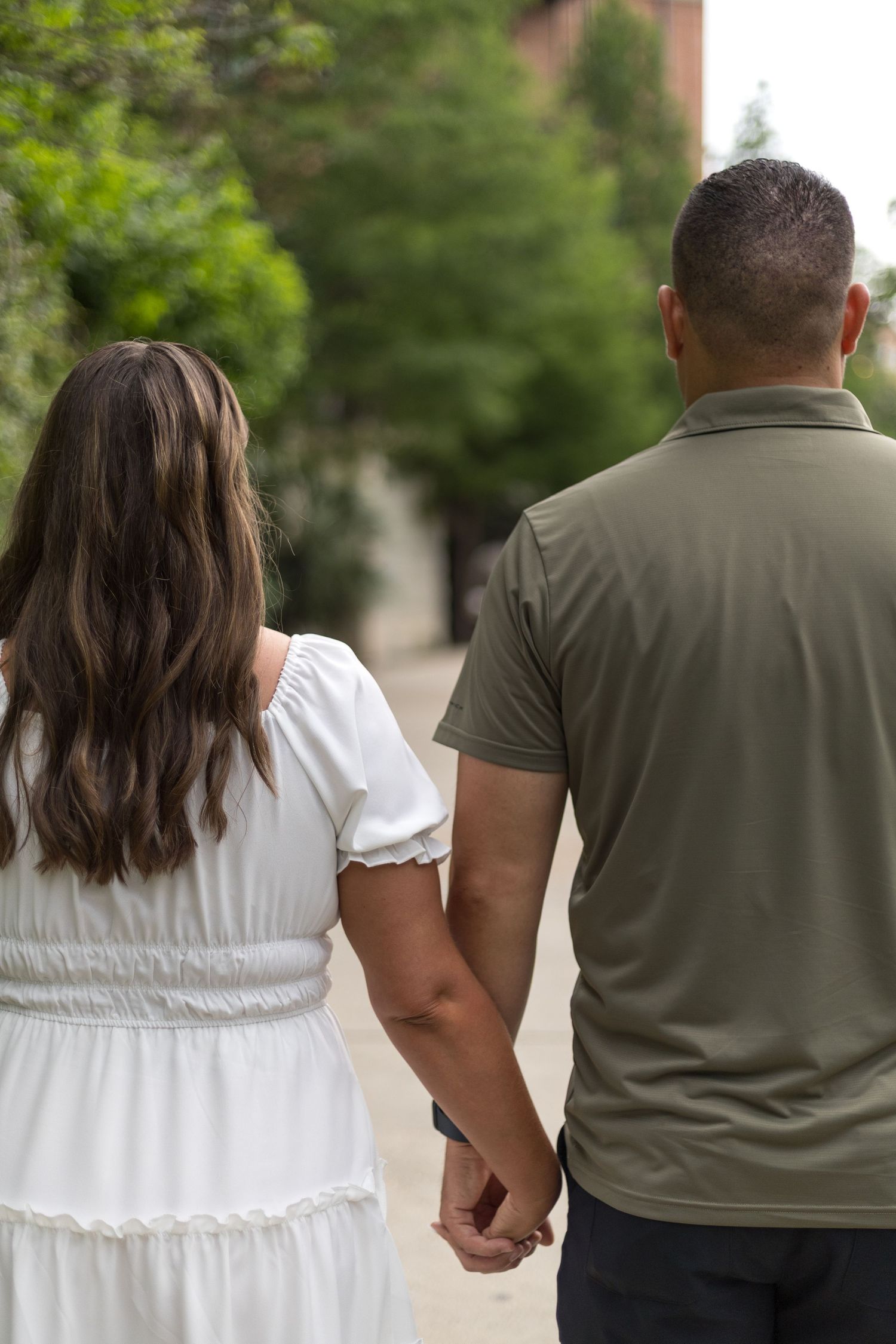 Engaged couple walking hand in hand down a tree-lined path, viewed from behind.