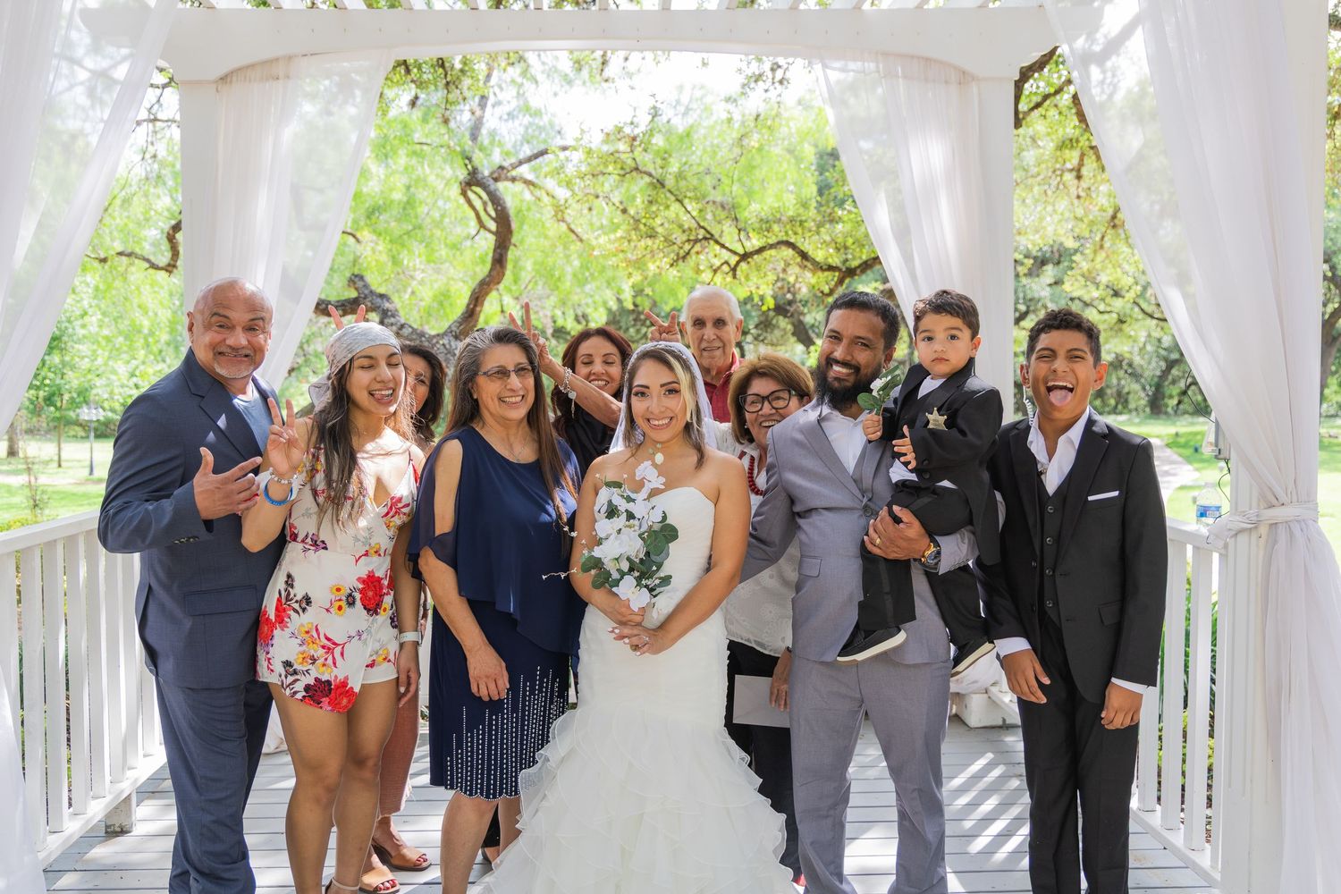 Bride and groom smiling with family members under a white draped gazebo outdoors.
