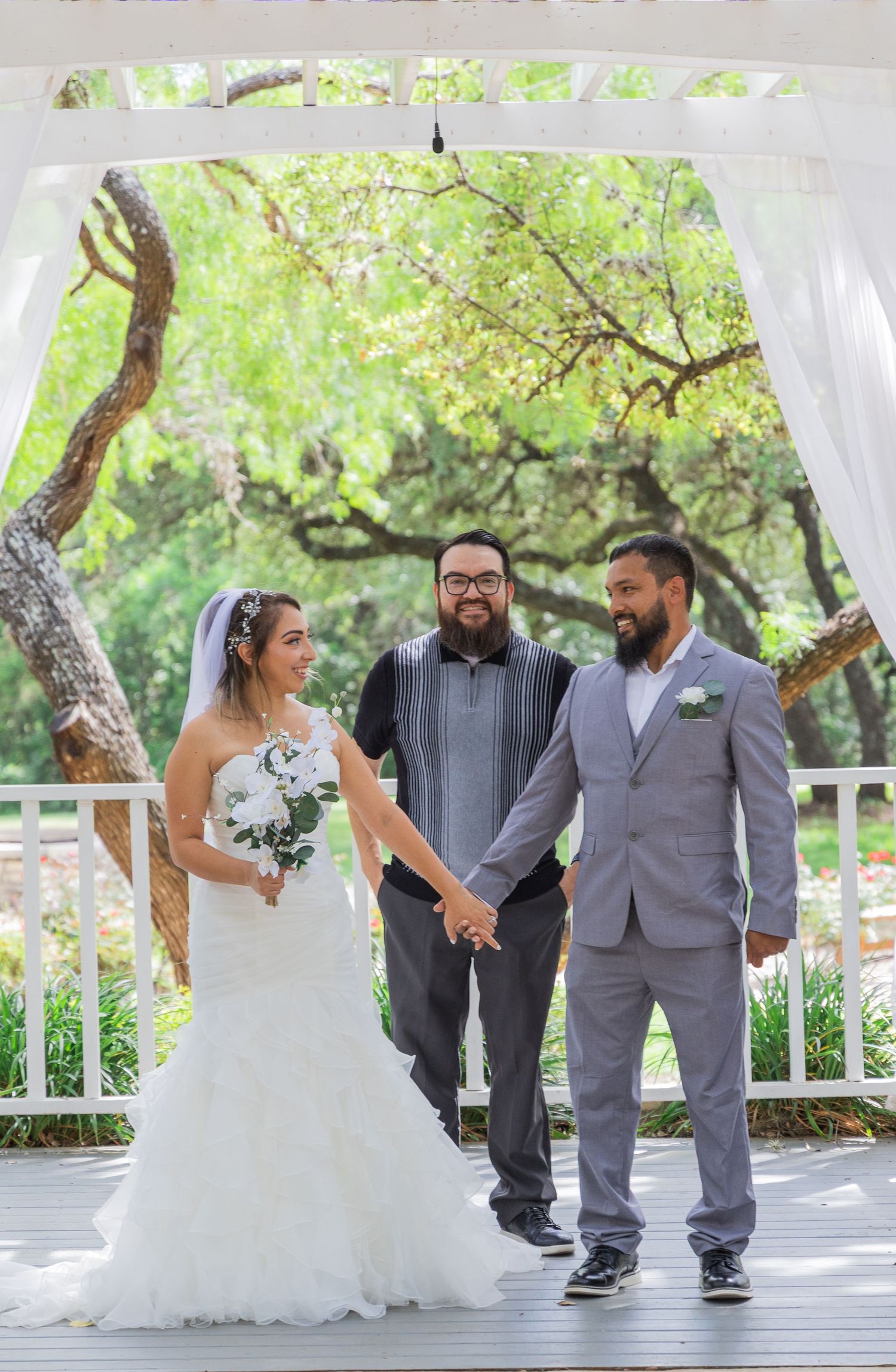 Bride and groom hold hands with smiling officiant under white pergola surrounded by lush greenery.
