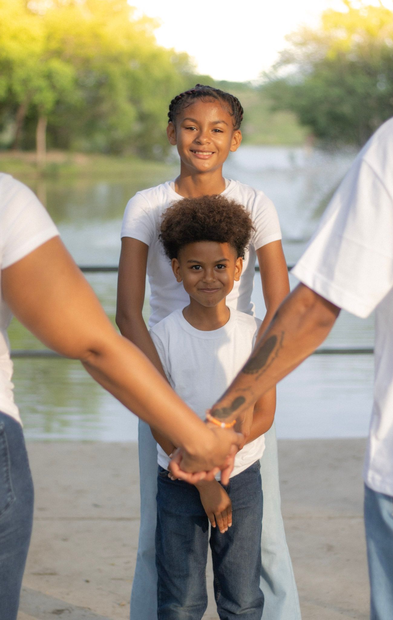 Two children in white shirts smile while parents hold hands in the foreground near a pond.