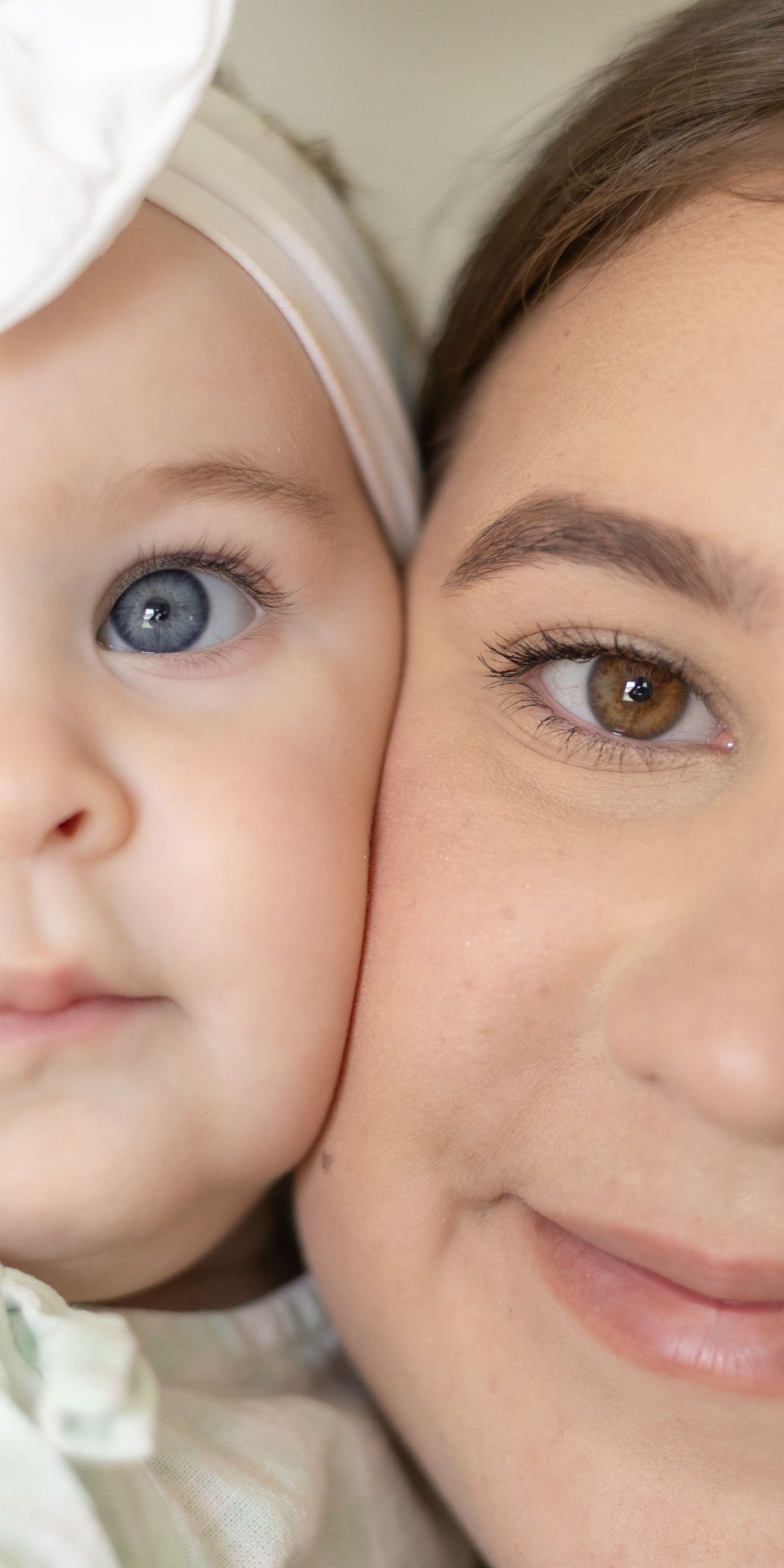 Close-up portrait of smiling mother and blue-eyed baby girl with white bow headband cheek to cheek.