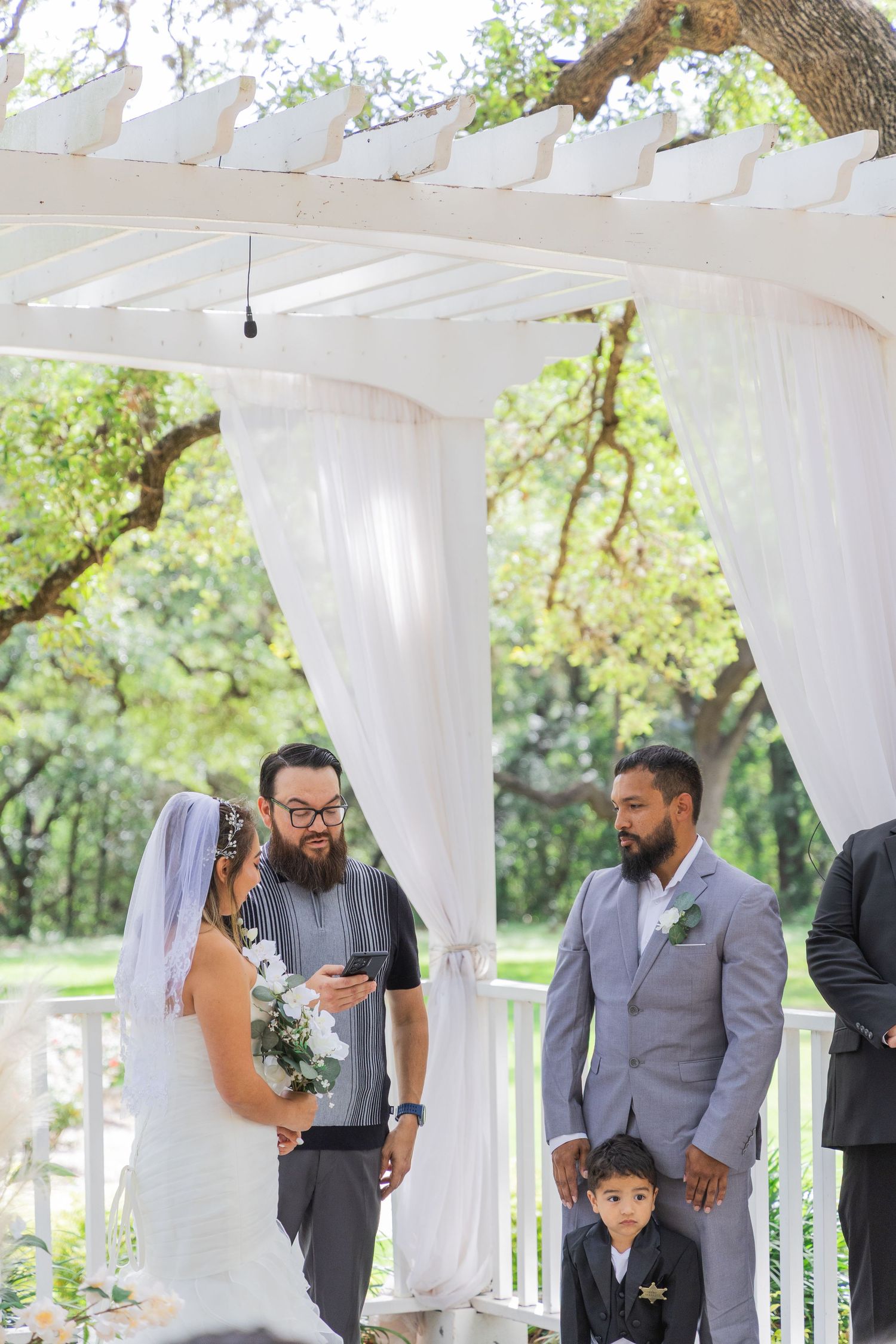 Officiant reads vows as bride, groom in gray suit, and ring bearer stand under white pergola.