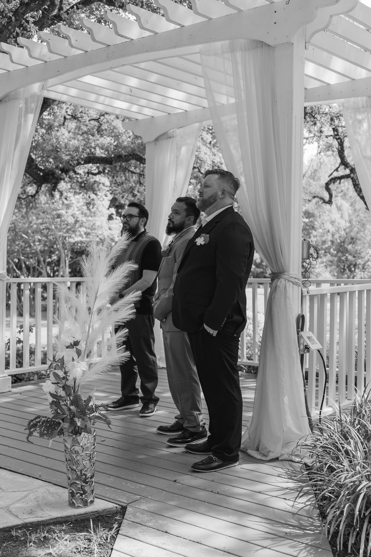 Groom and groomsmen stand at outdoor wedding altar under white draped pergola awaiting bride.