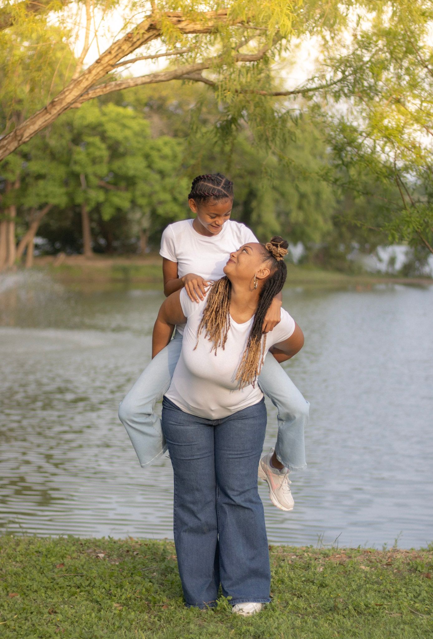 Joyful mother giving her young child a piggyback ride near a scenic pond with green trees.
