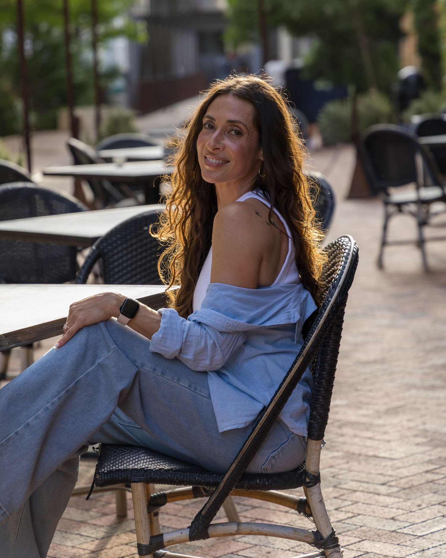 Woman in blue shirt and flare jeans sits relaxed in outdoor patio chair, smiling in warm sunlight.