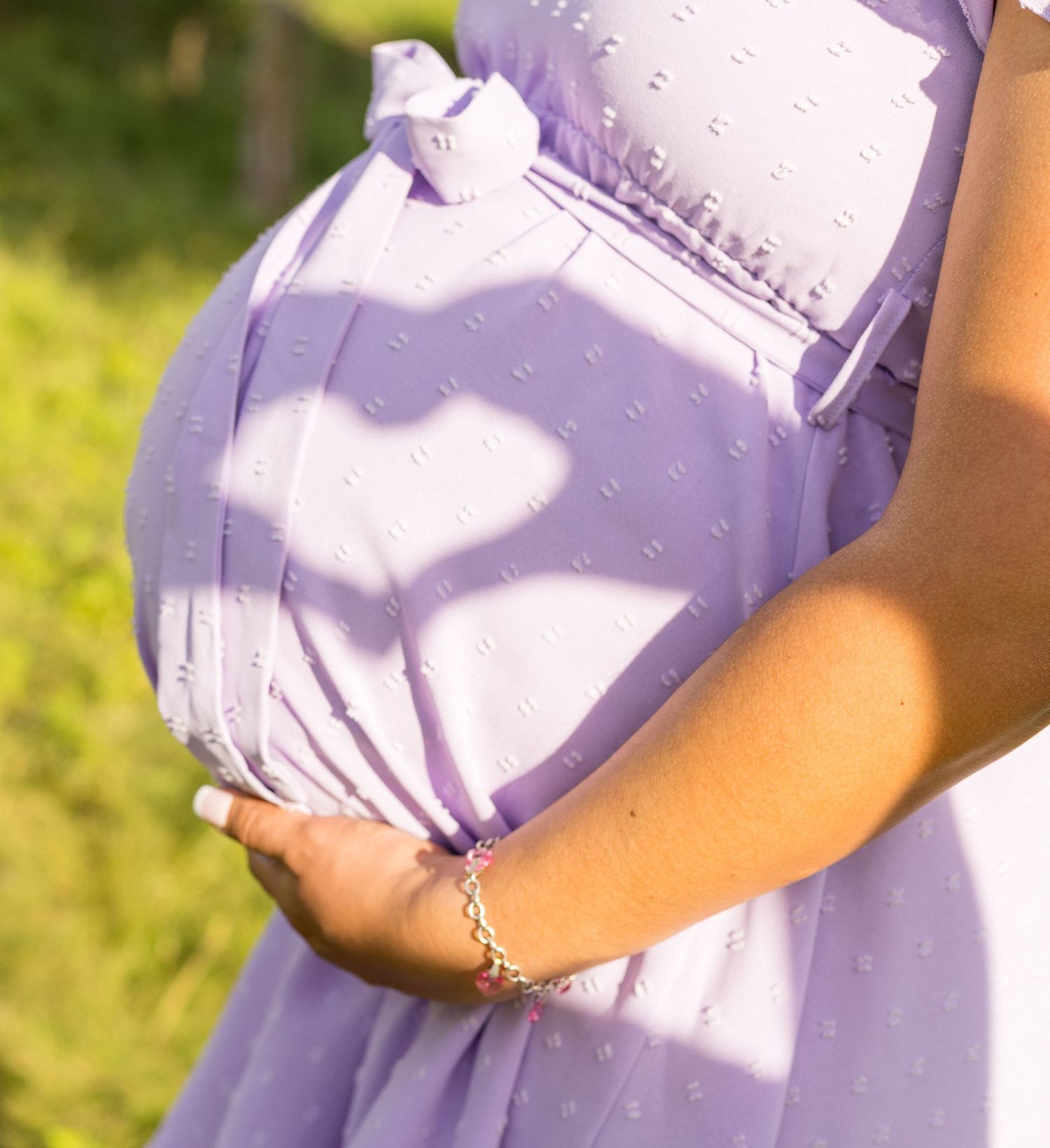 Close-up of pregnant woman in lavender dress gently holding baby bump in sunlit garden.