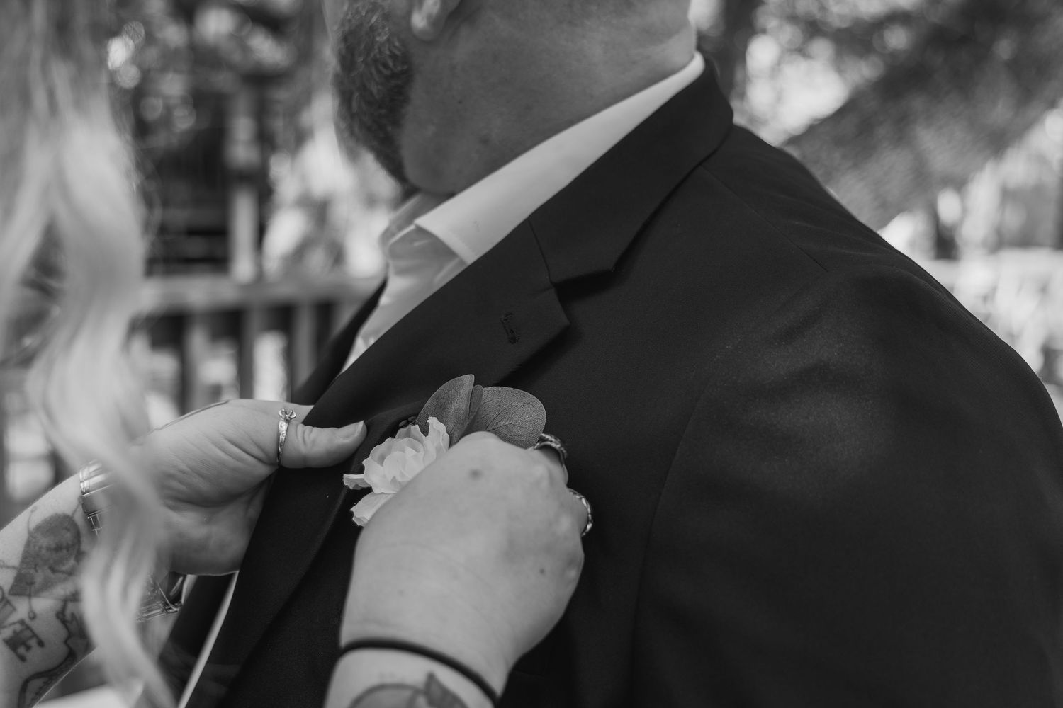 Black and white close-up of hands pinning boutonniere on groom in dark suit jacket.