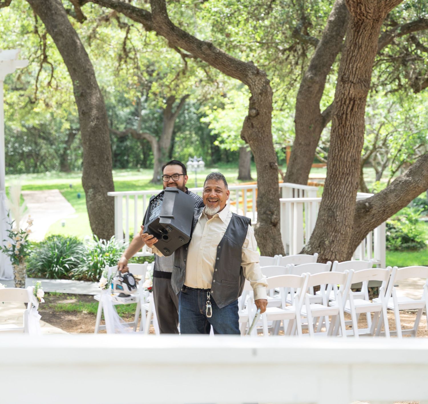 Two men smiling and posing at an outdoor wedding ceremony venue surrounded by oak trees and white chairs.