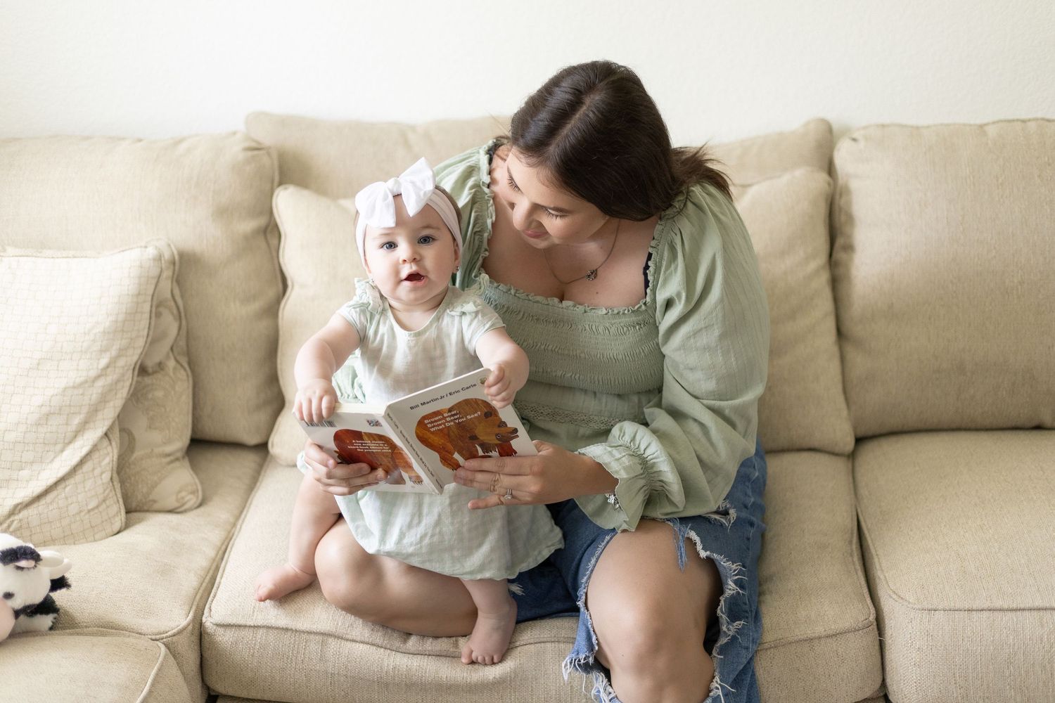 Mother in green top reading board book with baby girl in white bow headband on couch.