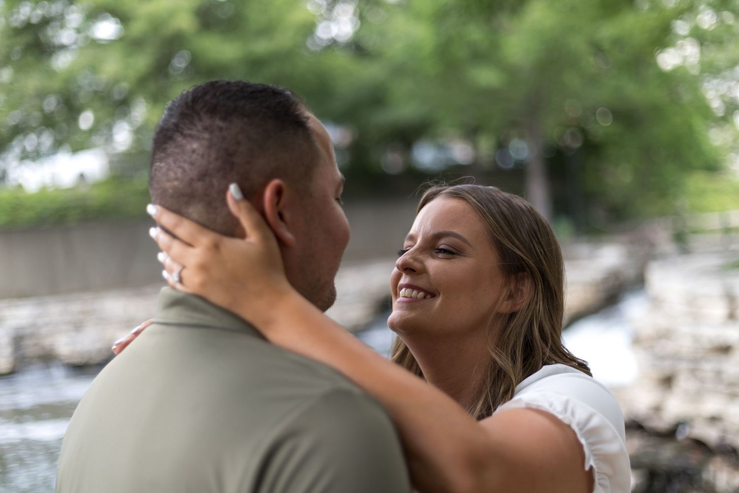 Happy woman smiling at her partner outdoors near a waterway surrounded by lush green trees.