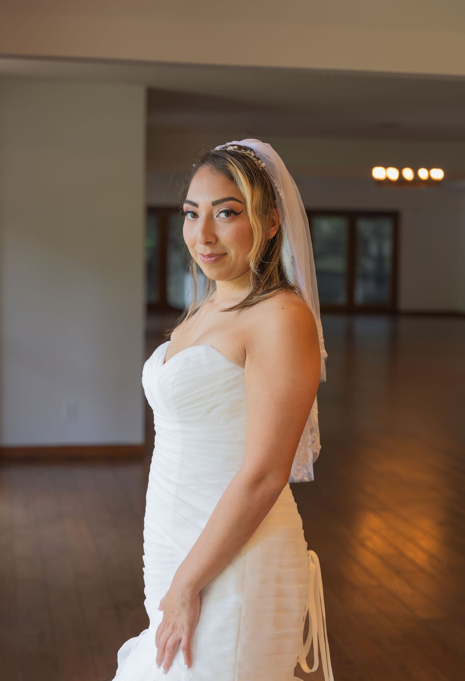 Bride in a strapless ruched wedding gown with veil posing elegantly in a sunlit reception hall.