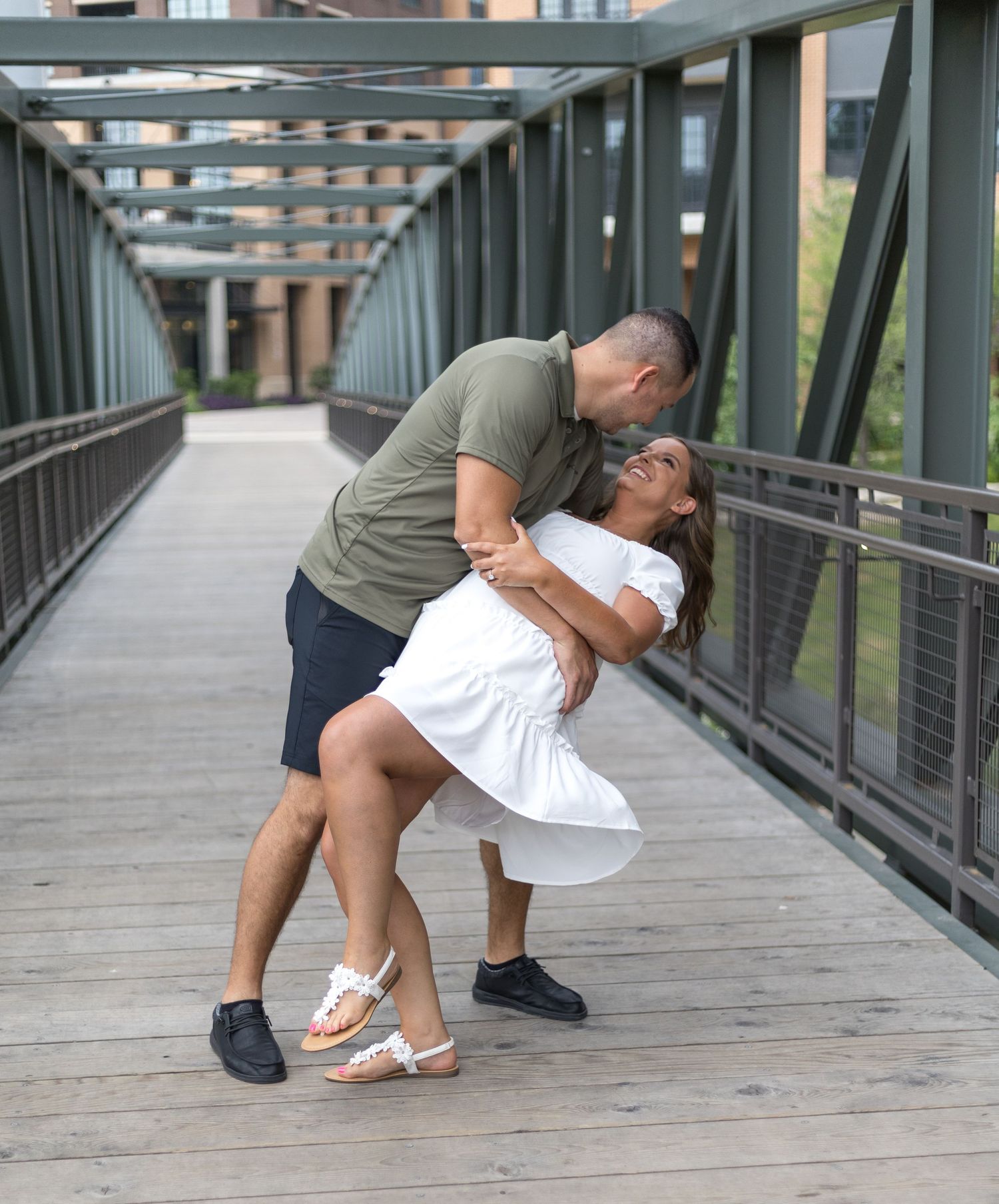 Couple sharing a romantic dip kiss on a wooden bridge walkway with urban architecture in the background.