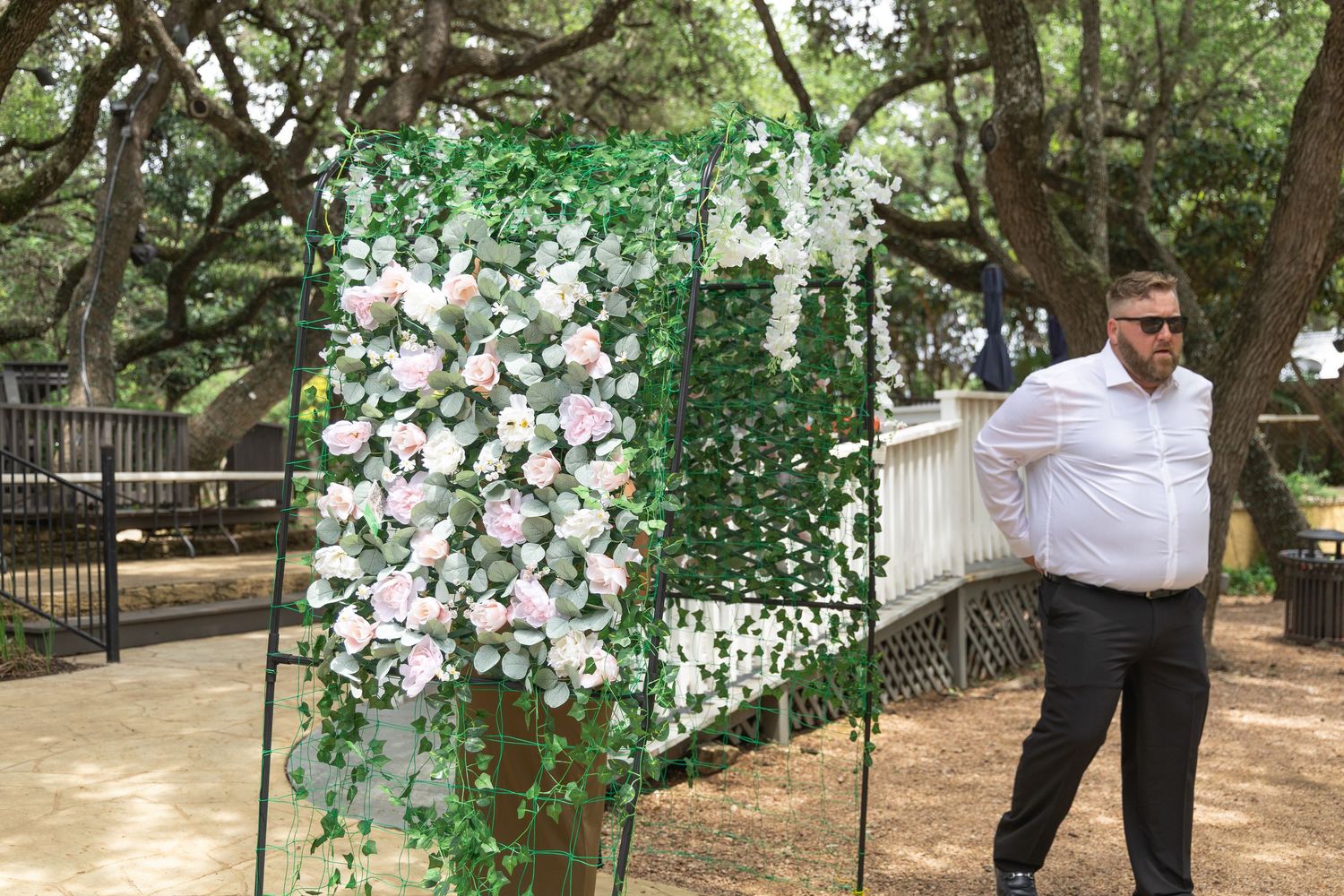 Man in white shirt standing beside a floral wall backdrop with pink roses at an outdoor wedding venue.