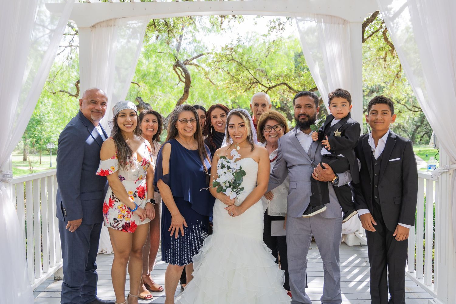 Wedding party group photo under white gazebo with bride and groom surrounded by family.
