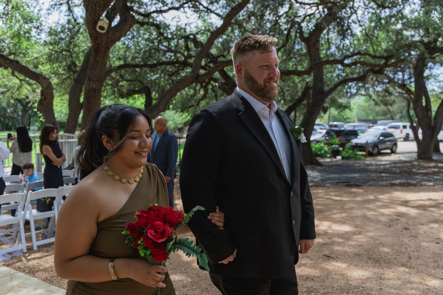 Woman in gold dress holds red roses while walking with bearded man in black suit at outdoor wedding.