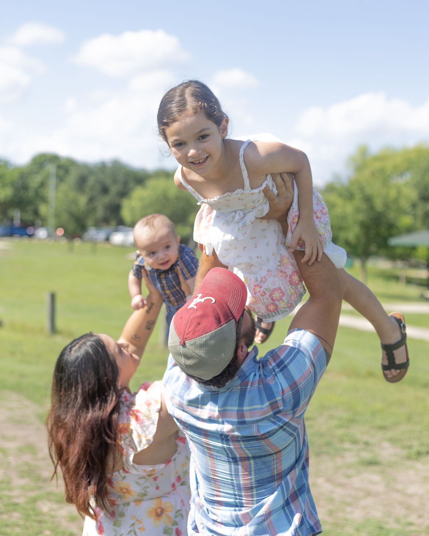 Joyful father lifts smiling daughter in white dress while mother holds baby boy in sunny park.