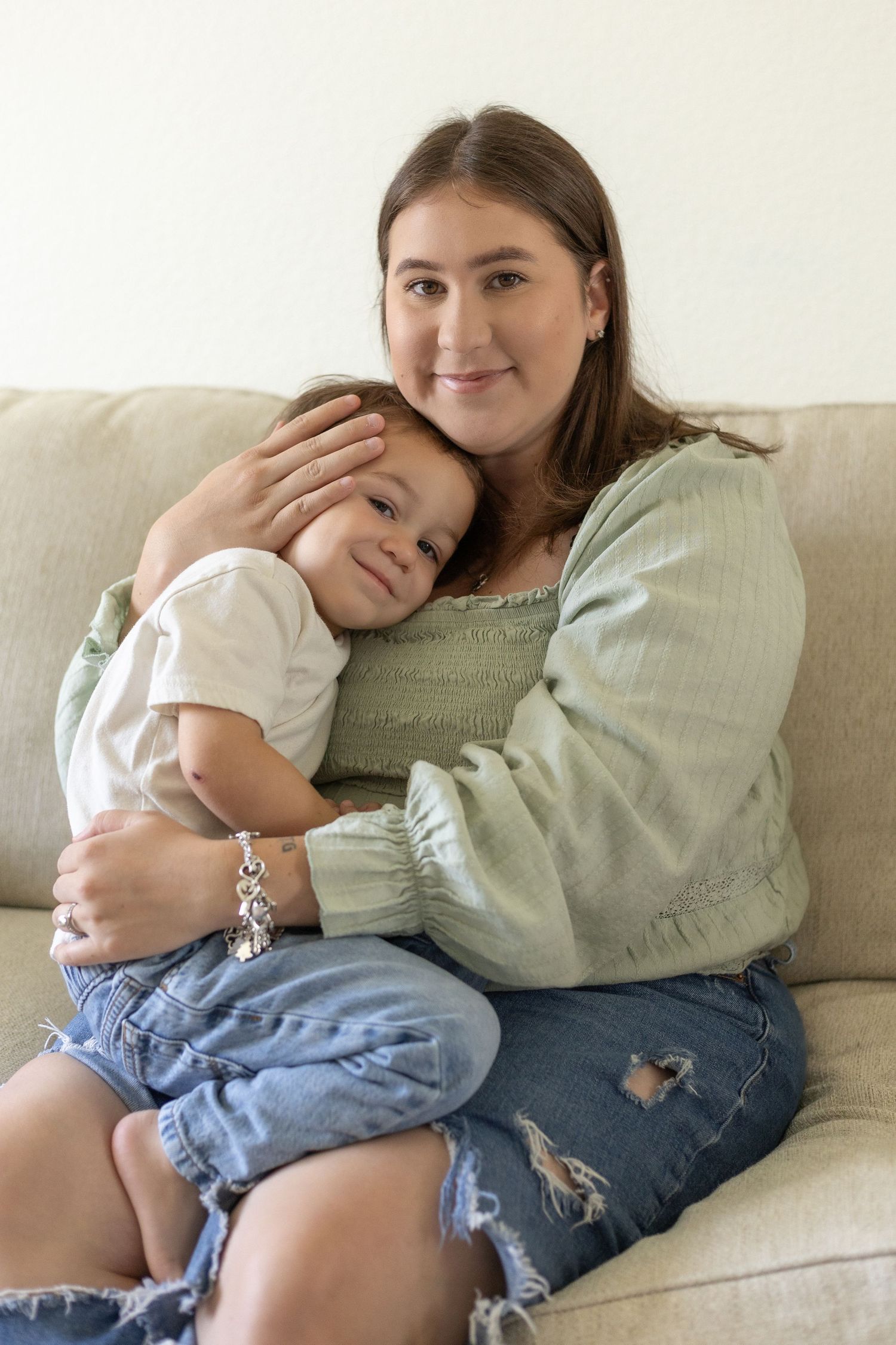 Young mother in green top hugging toddler son on beige sofa at home, smiling at camera.