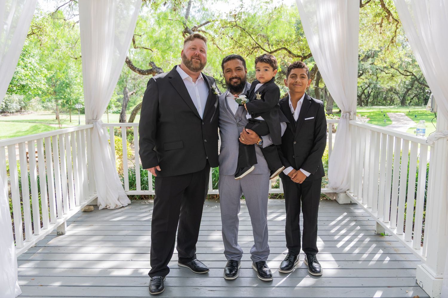 Three groomsmen in dark suits posing with young boy under white gazebo outdoors.