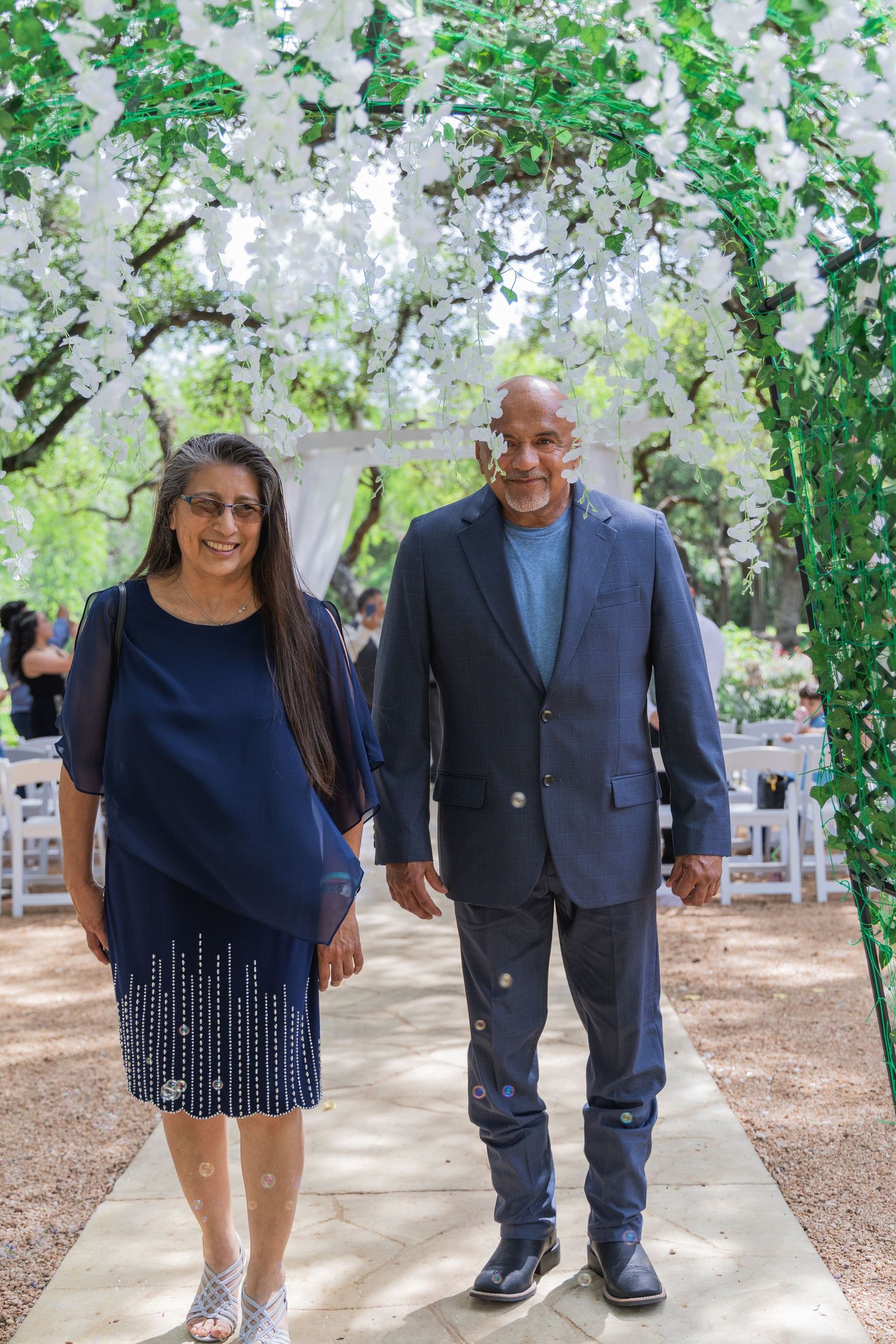 Couple in coordinating navy outfits walks together under white floral arch at outdoor wedding venue.