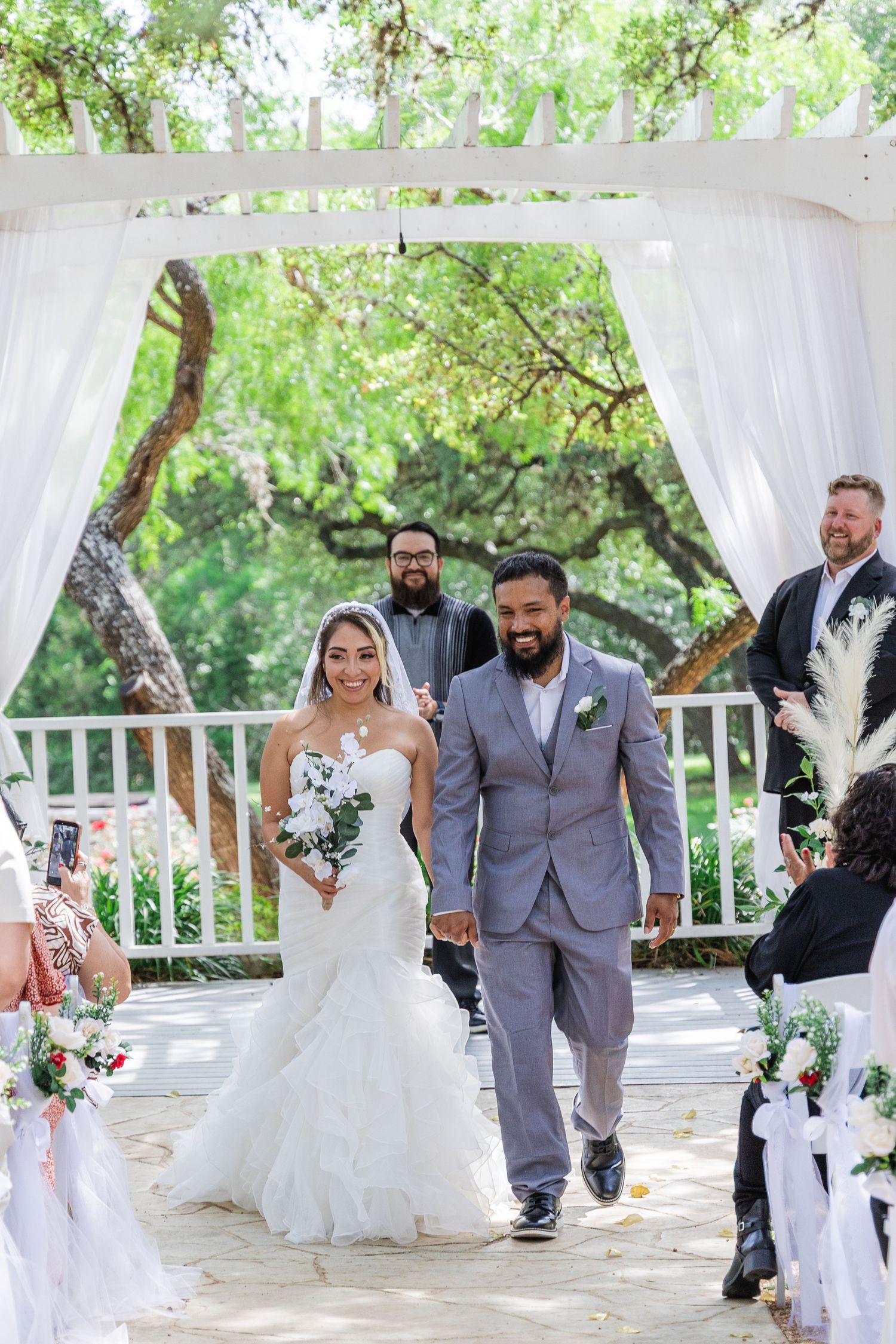 Newlyweds walk hand in hand down aisle after outdoor wedding ceremony under white draped arbor.