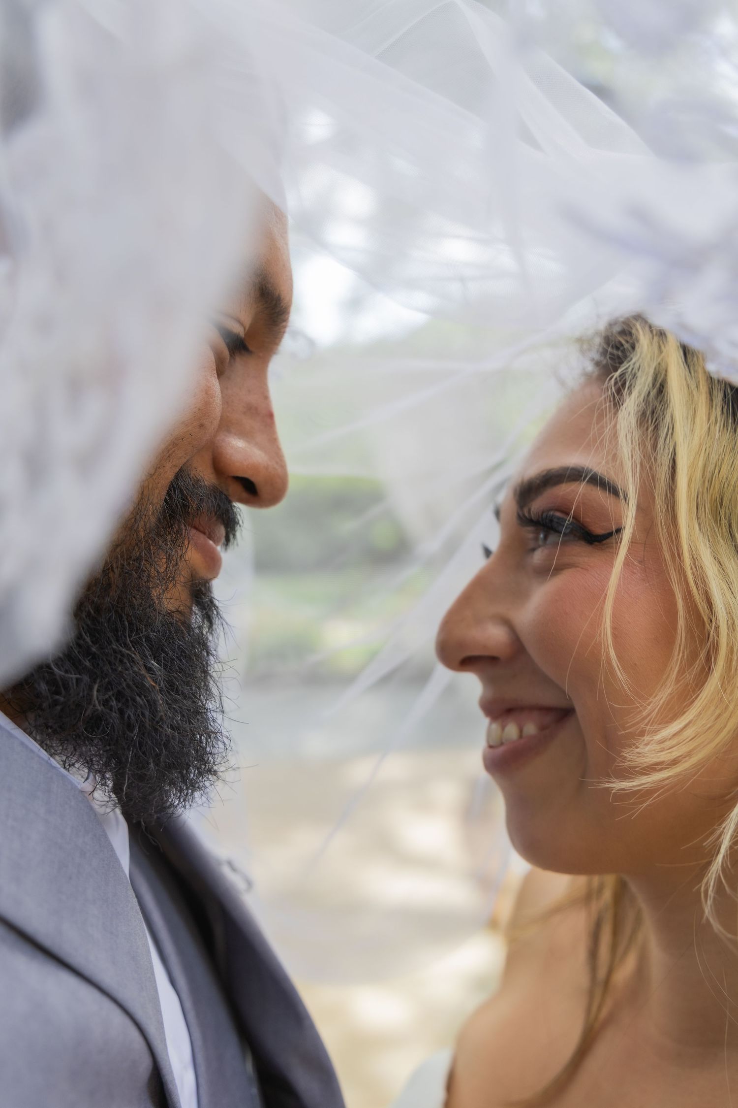 Close-up of bearded groom and blonde bride smiling at each other under a flowing white wedding veil.