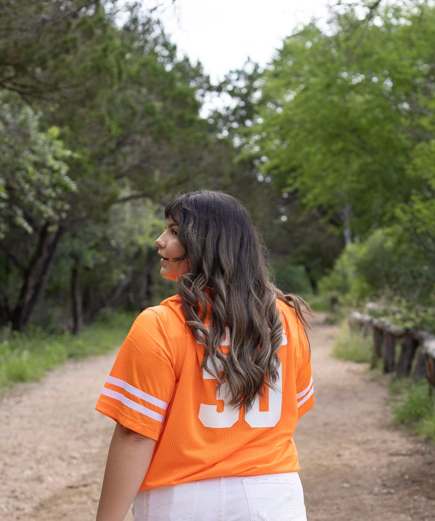 Back view of woman in orange UTRGV jersey walking on tree-lined dirt path.
