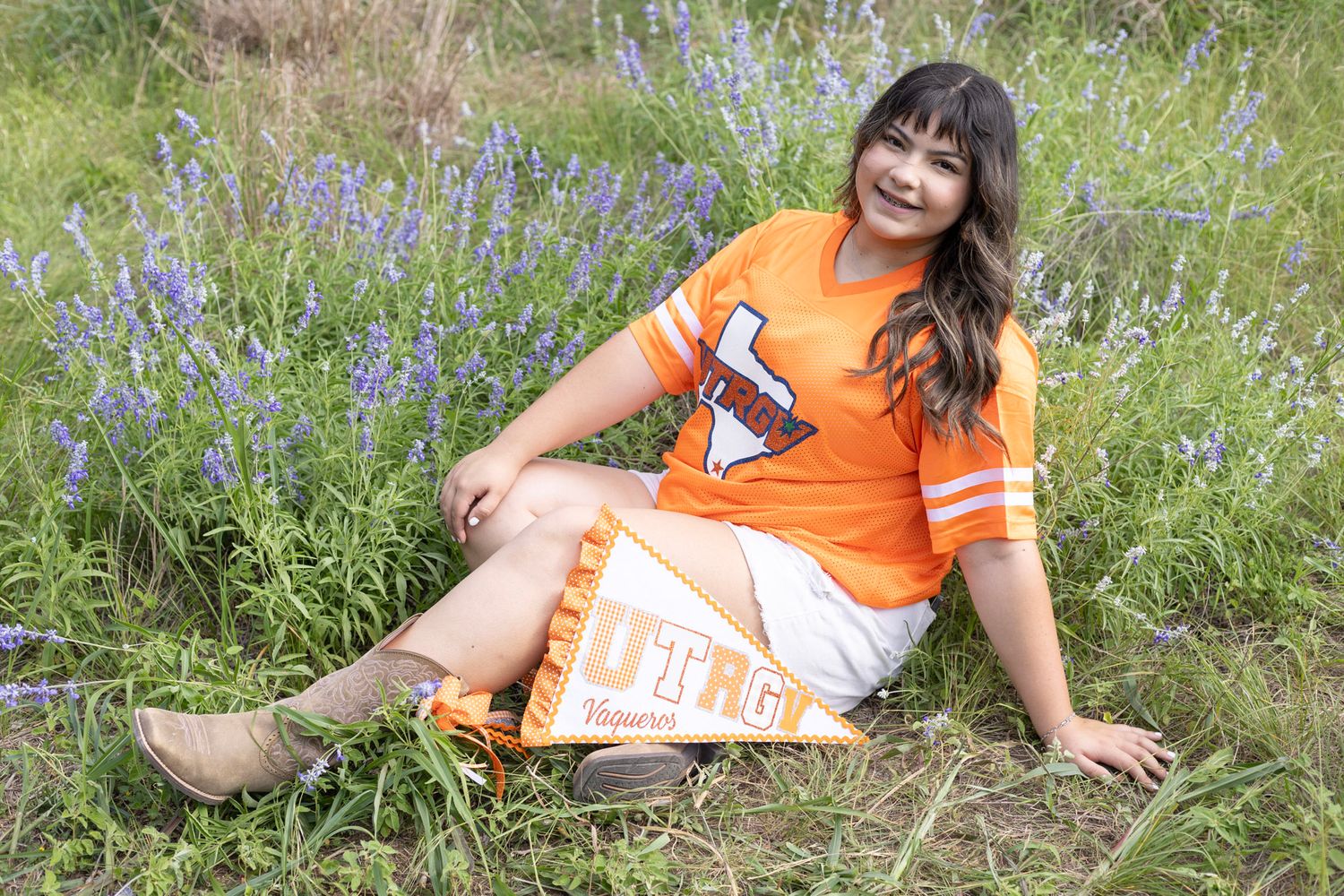 Young woman in orange UTRGV jersey sits smiling in vibrant bluebonnet field.