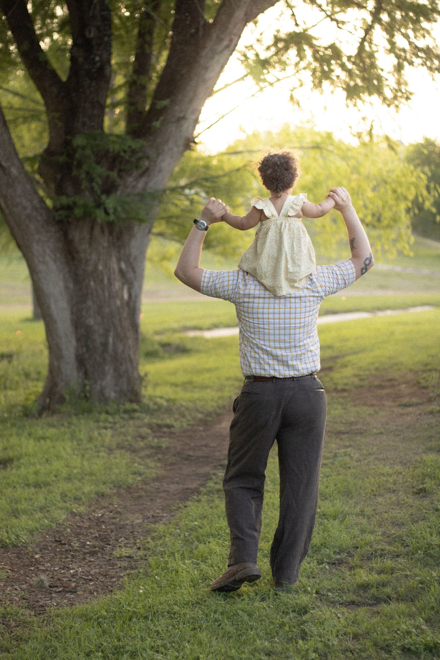 A father carries his young daughter on his shoulders while walking through a lush green park near a large tree.