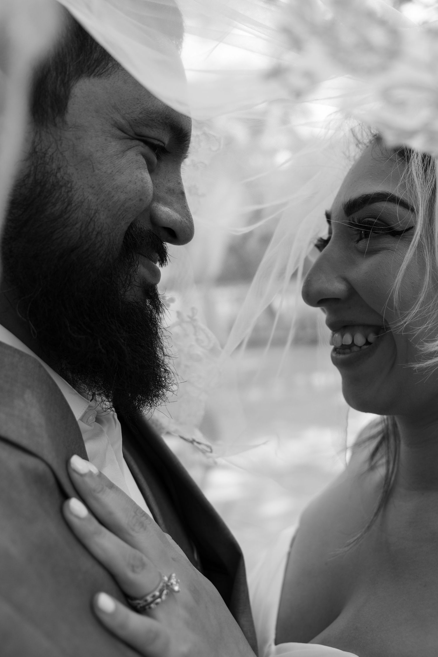 Black and white portrait of laughing bride and bearded groom sharing a joyful moment under wedding veil.