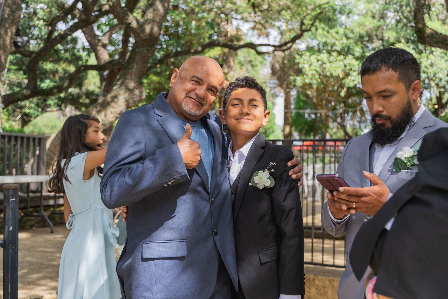 Man in blue suit poses with teen boy giving thumbs up at outdoor wedding celebration.