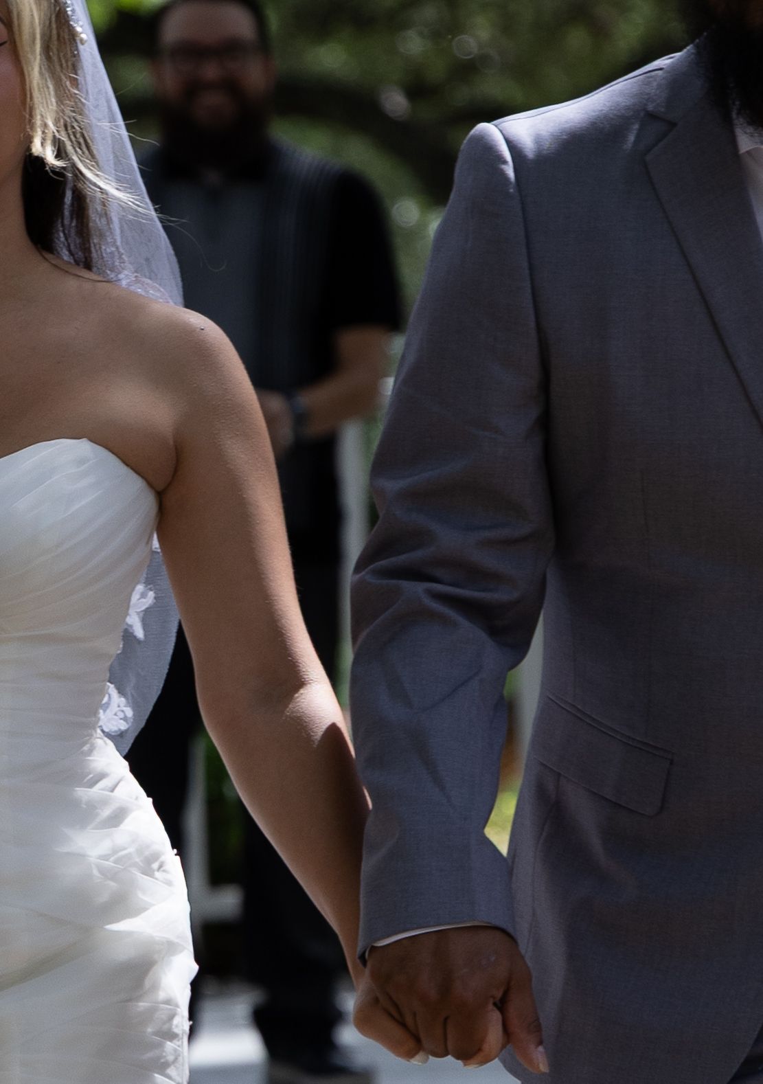 Close-up of bride and groom holding hands at outdoor wedding ceremony with officiant in background.