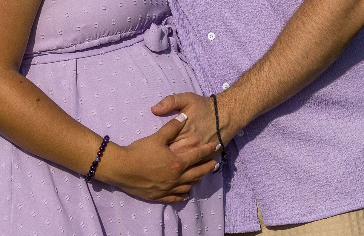 Close-up of couple holding hands over pregnant belly in matching lavender outfits outdoors.