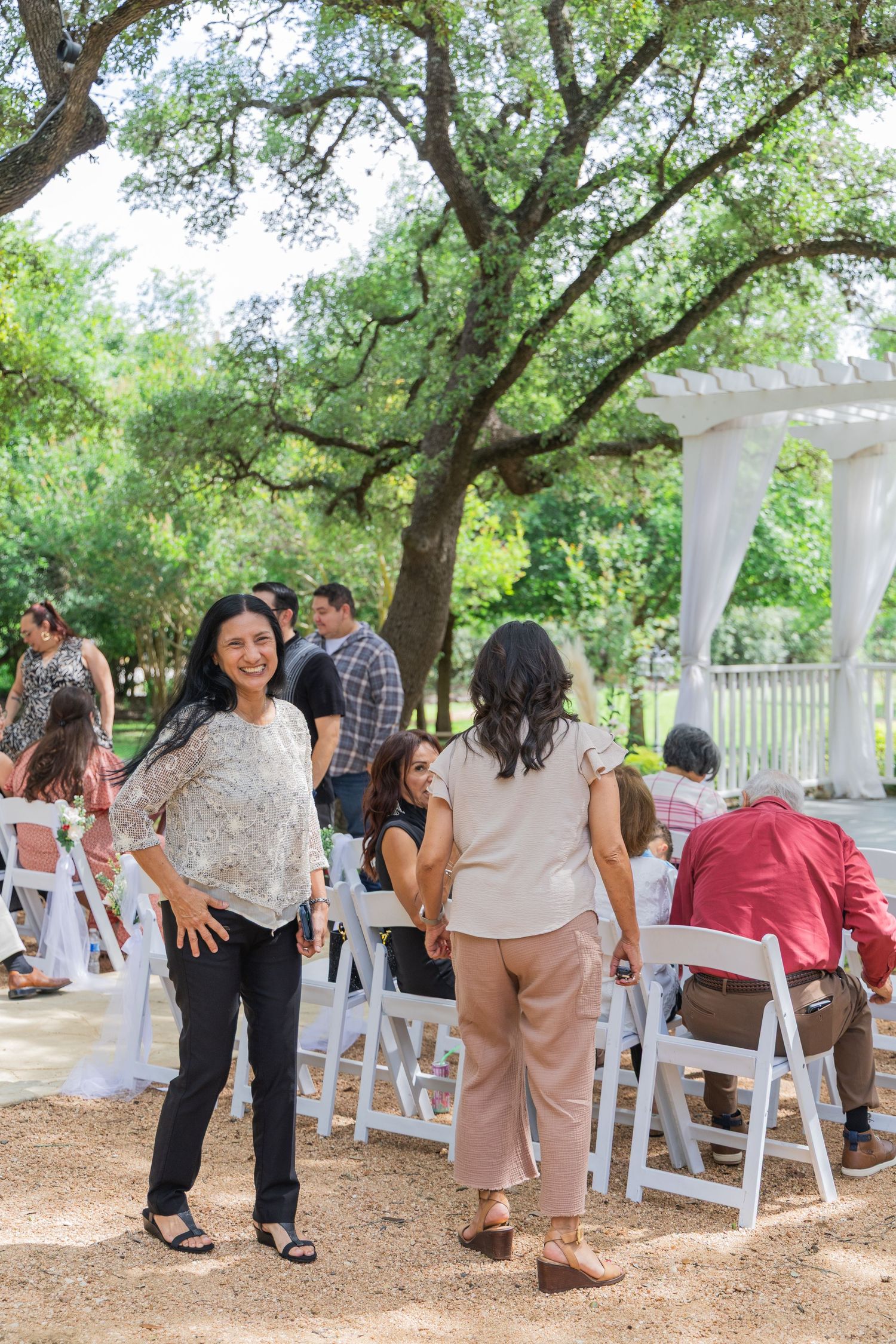 Smiling woman in floral top posing at outdoor wedding venue surrounded by guests and oak trees.