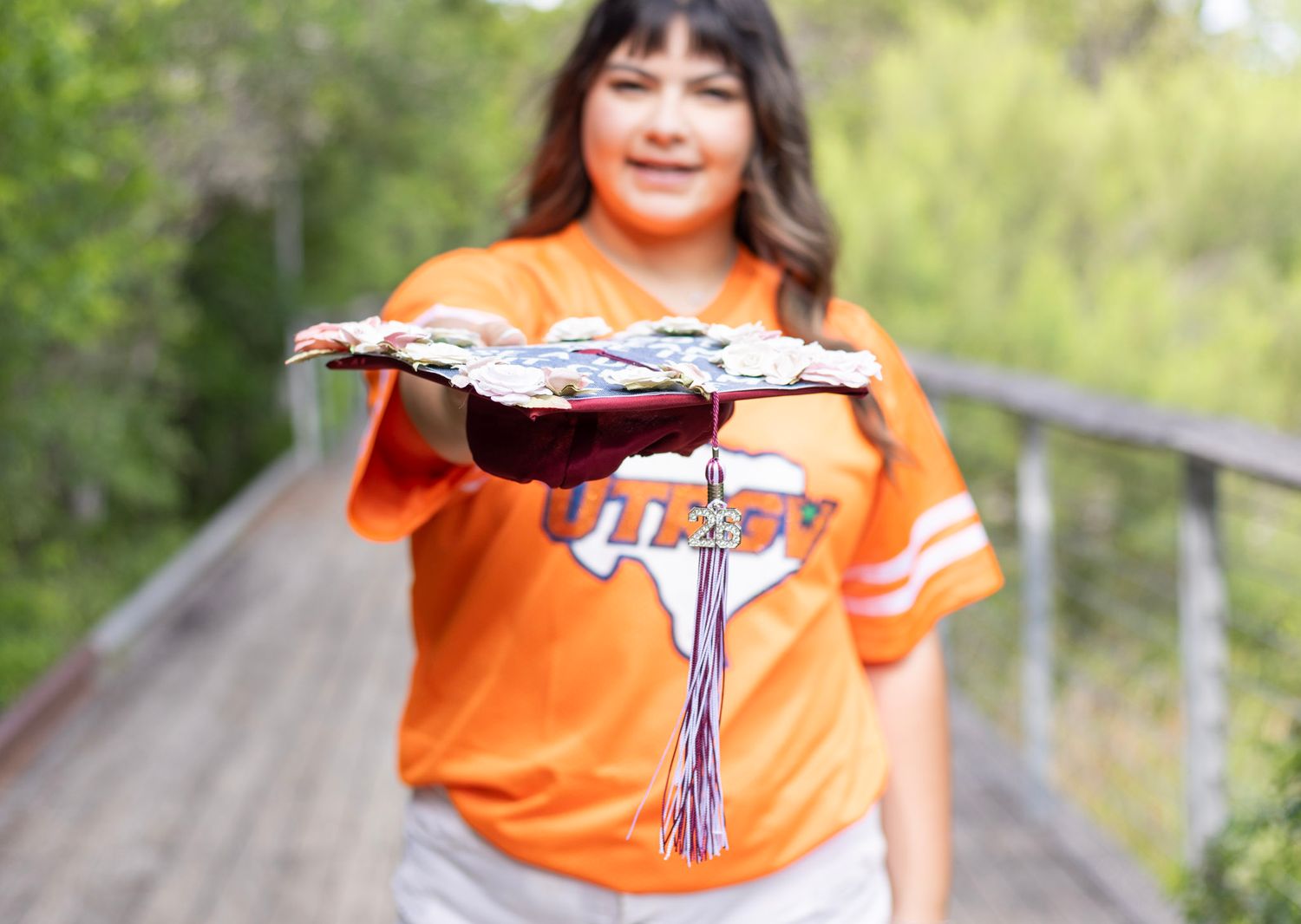 Graduate in orange Texas shirt holding decorated graduation cap toward camera on wooden bridge.
