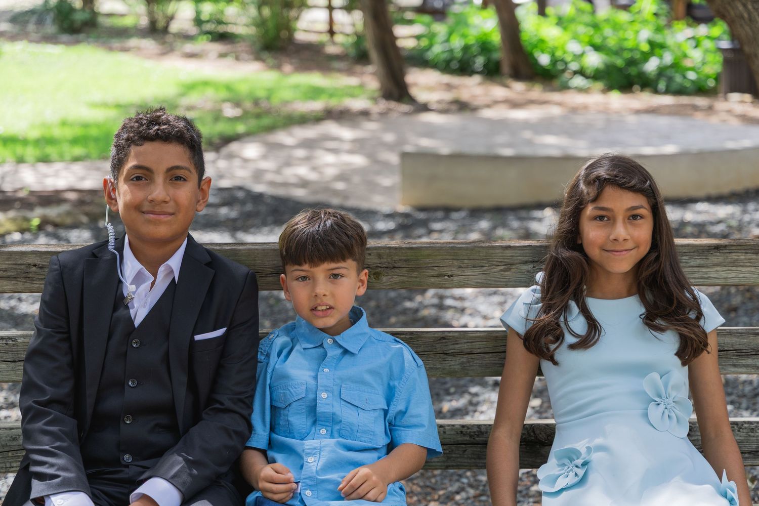 Three children dressed formally sitting on a bench in a park setting.