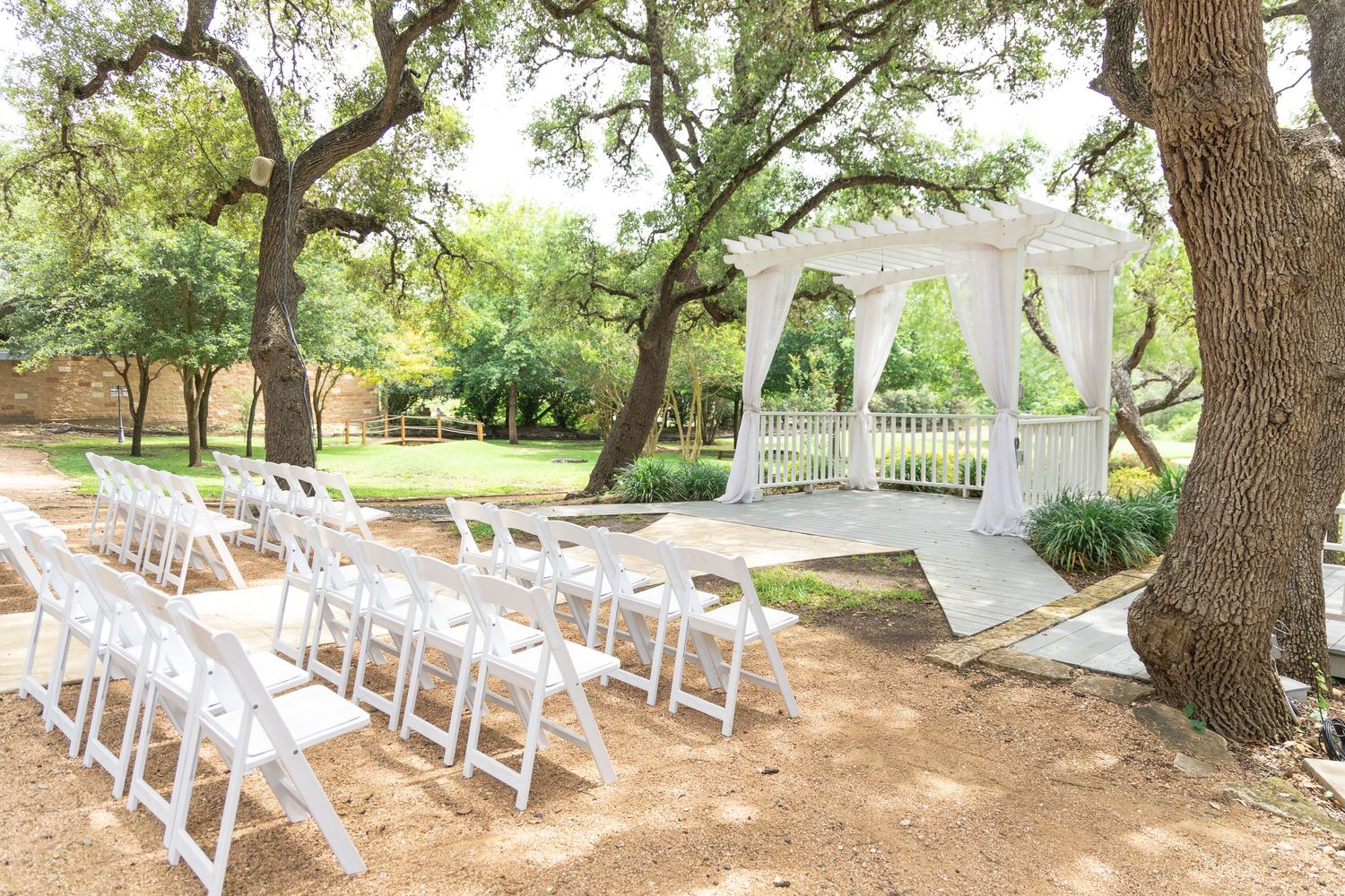 Sunlit outdoor wedding ceremony setup with white chairs, draped arbor, and mature oak trees in background.
