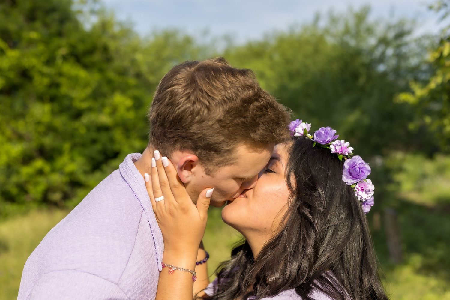 Couple sharing a kiss outdoors, woman wearing purple floral crown during maternity photoshoot.