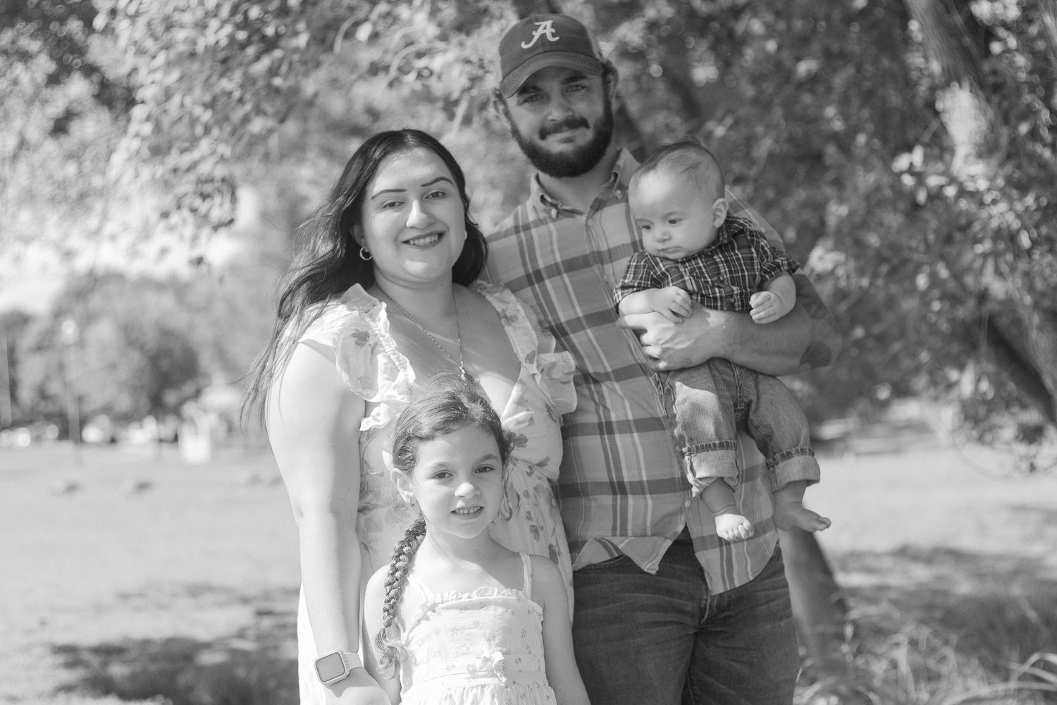 Black and white portrait of a loving family of four posing together under a shady tree outdoors.