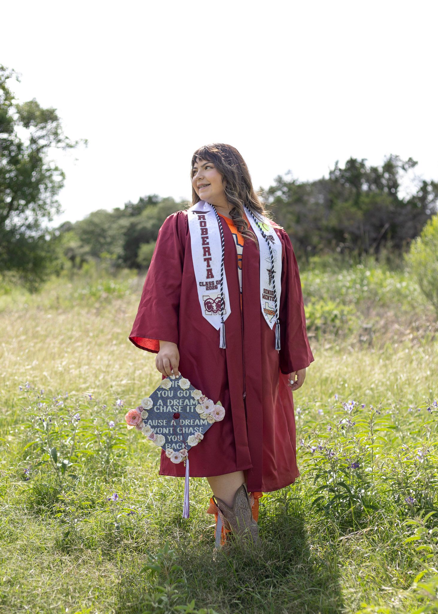 Smiling graduate in maroon gown holds decorated cap in lush green meadow.