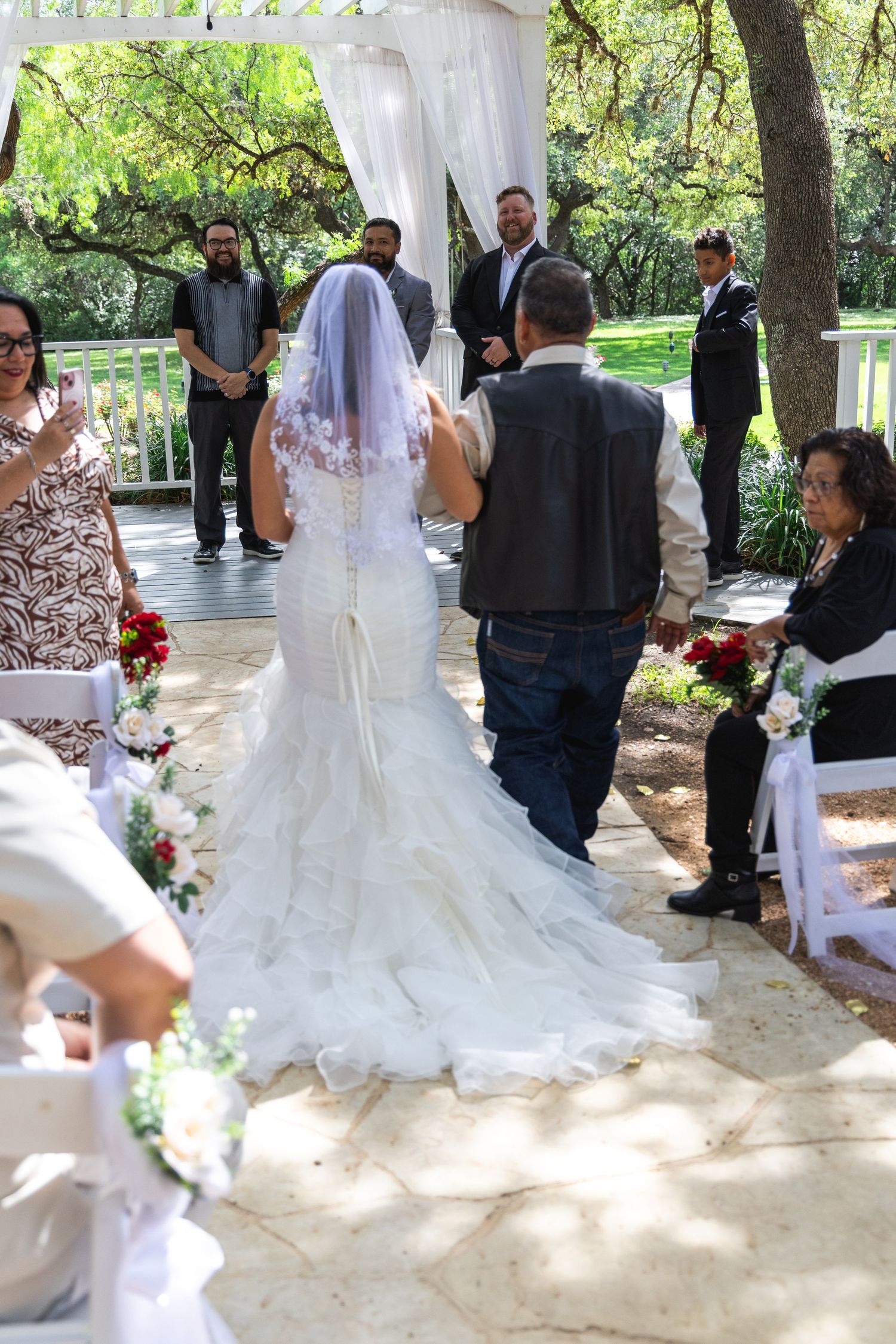 Bride in mermaid gown with veil walks down aisle with escort at outdoor garden wedding ceremony.