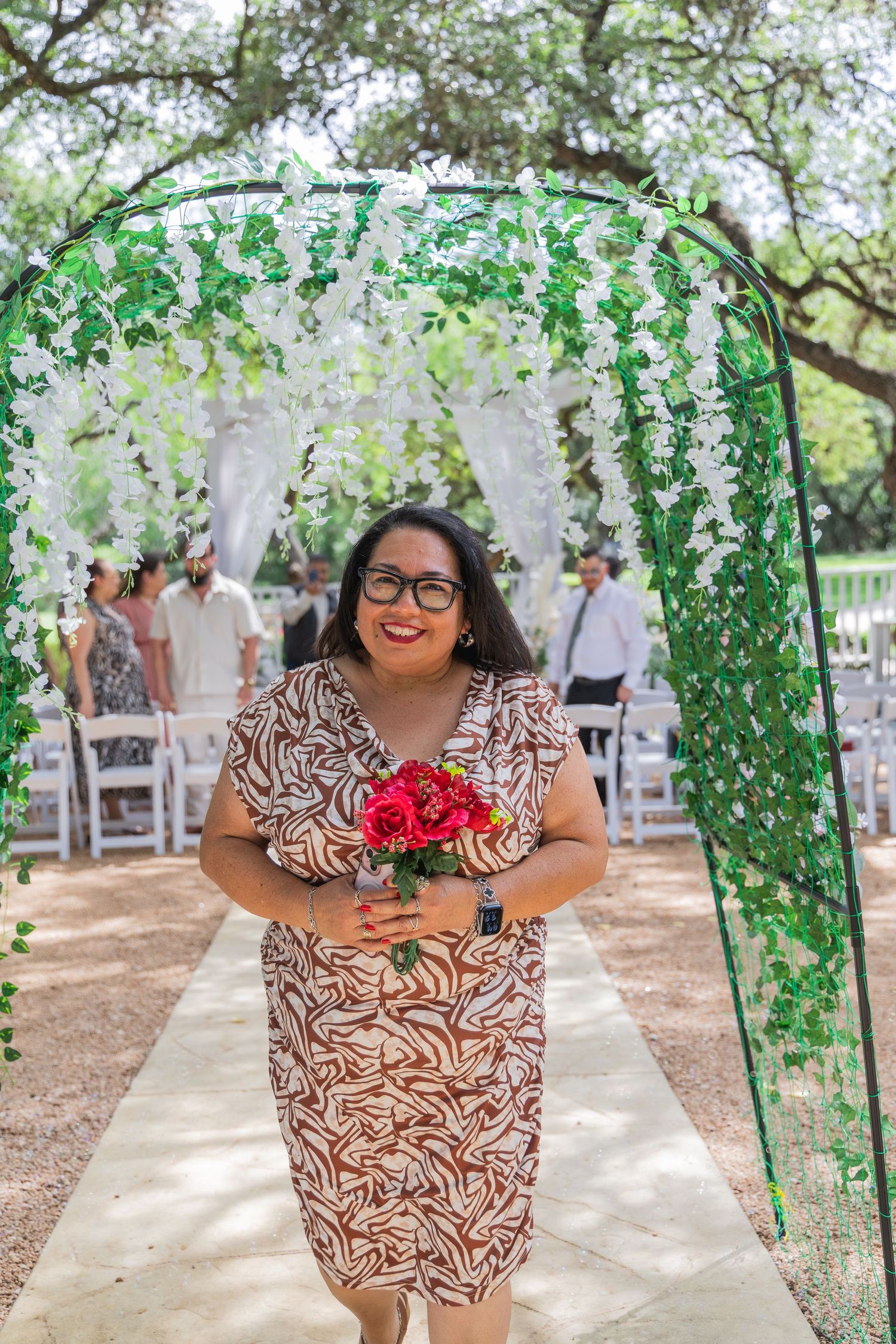 Smiling woman in patterned dress holds red roses under floral arch at outdoor wedding ceremony.