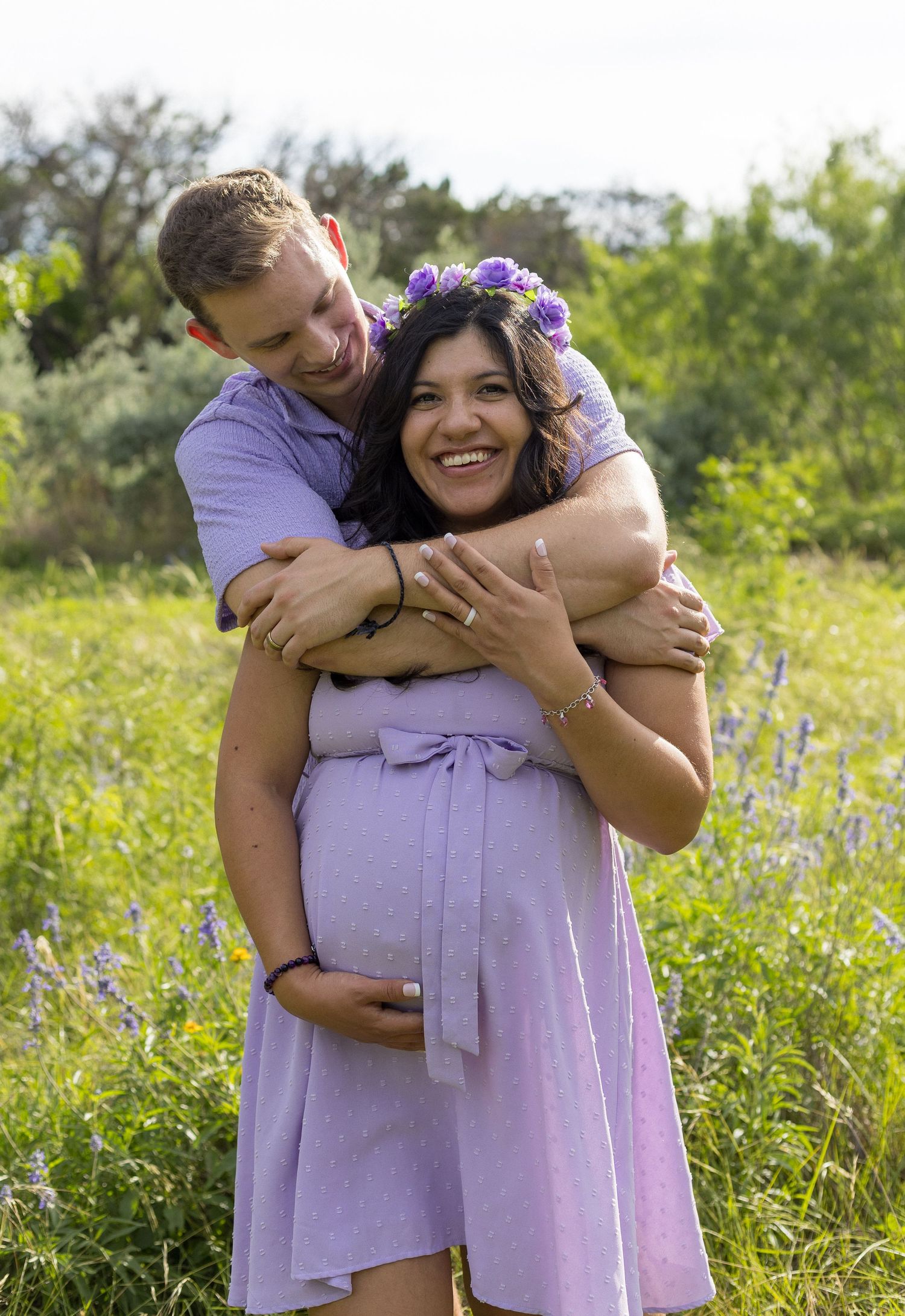 Happy pregnant woman in lavender dress with floral crown laughs as partner embraces her in green field.