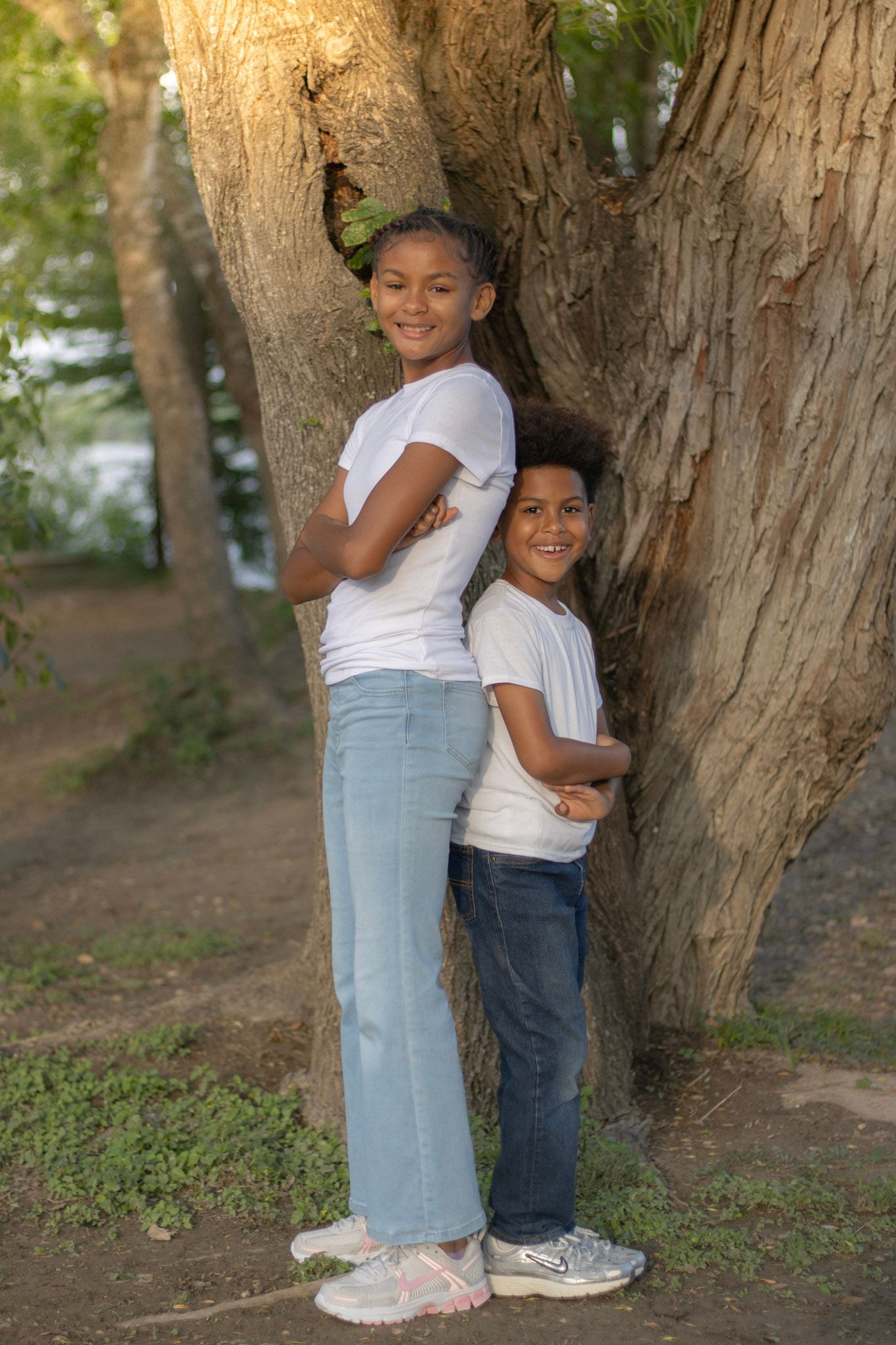Two children in matching white tees and jeans pose back-to-back against a large tree in a park.