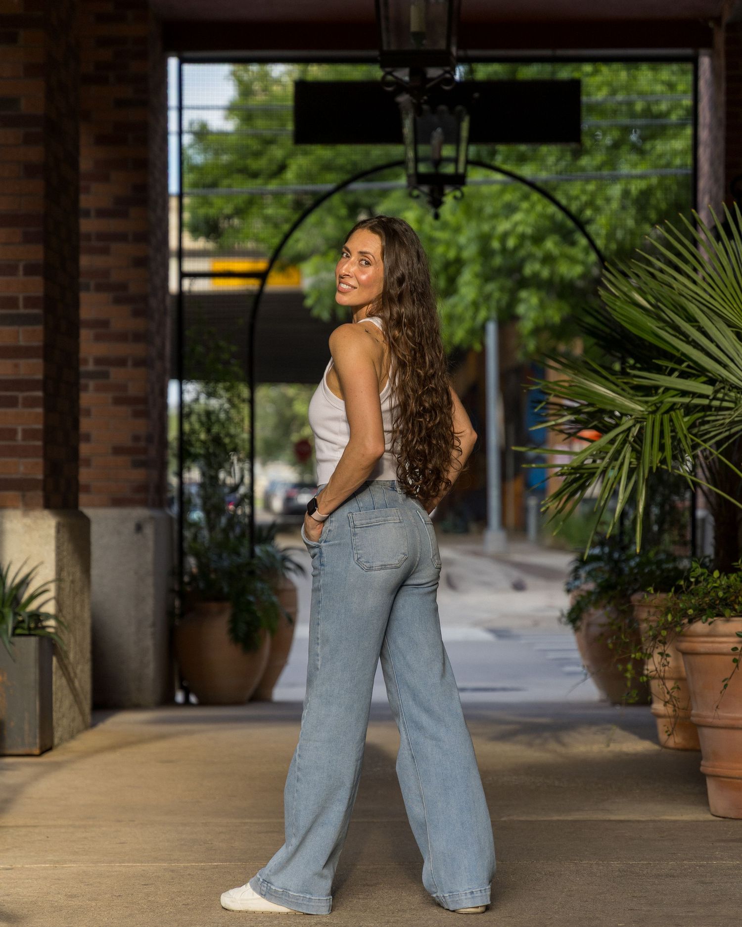 Brunette woman in white tank and wide-leg jeans poses gracefully under an arched entrance with tropical plants.