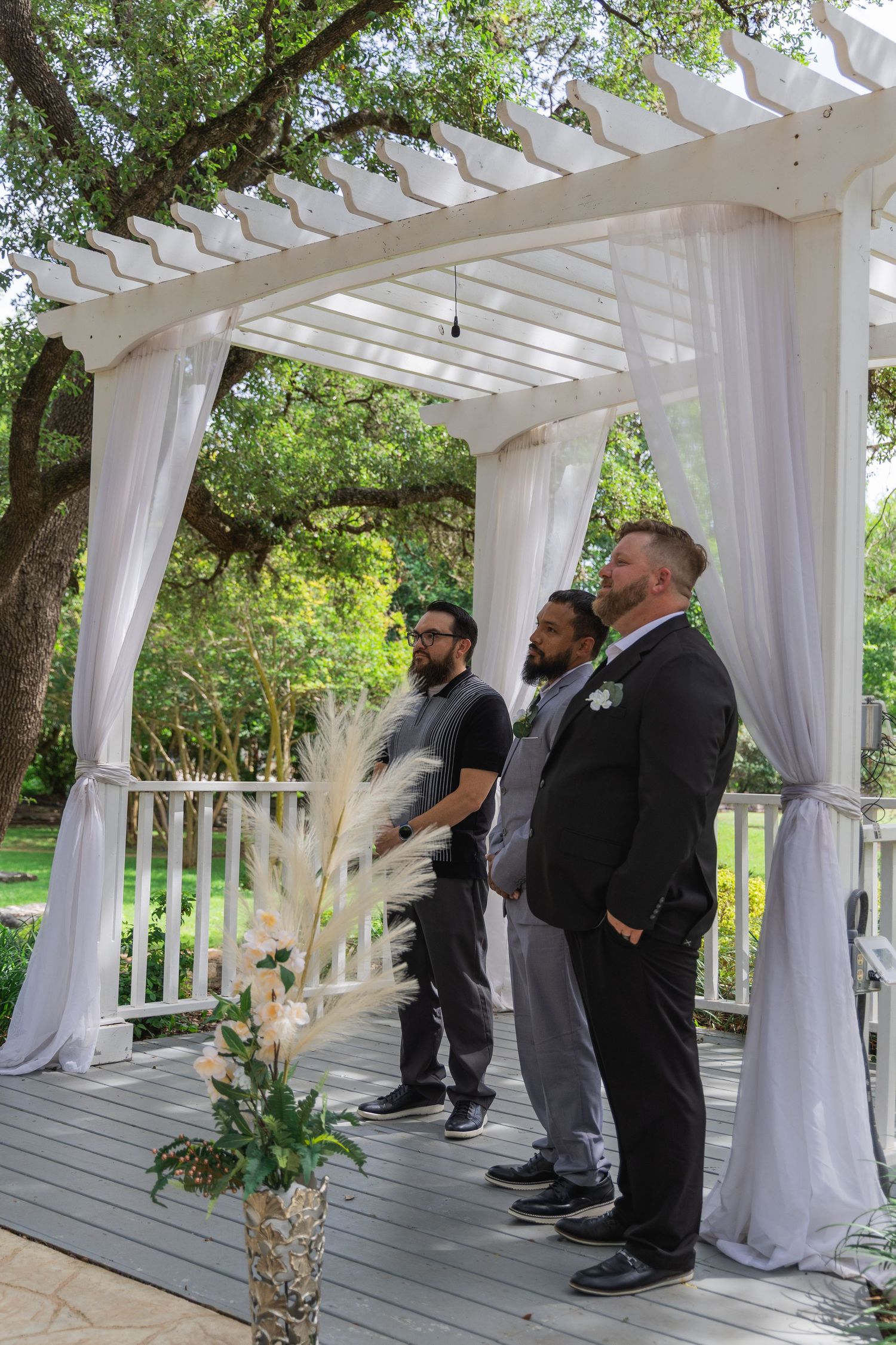 Groom in black suit standing with two groomsmen under white draped pergola at outdoor wedding.